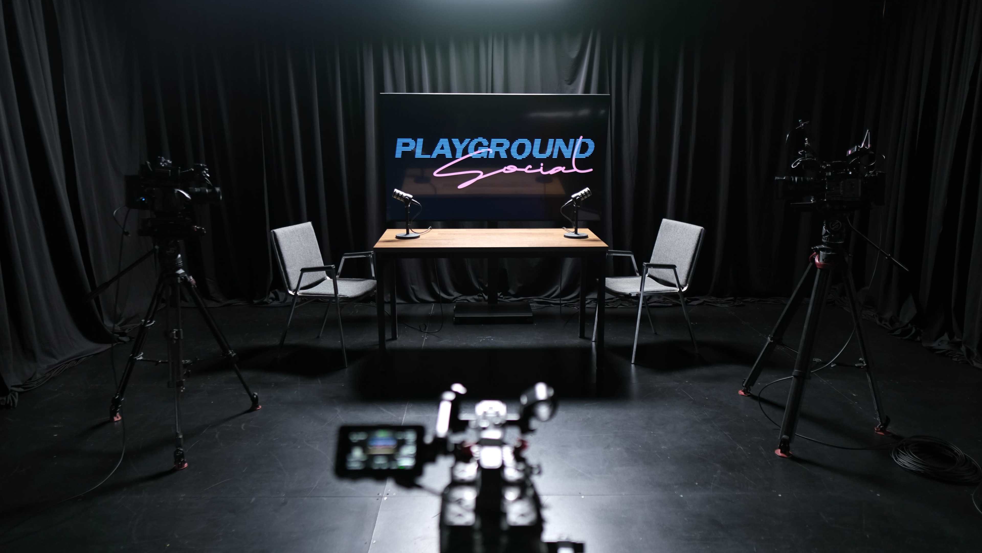 The image shows a TV studio setup with two chairs and a table, featuring a screen displaying "PLAYGROUND Social" and cameras positioned on tripods.