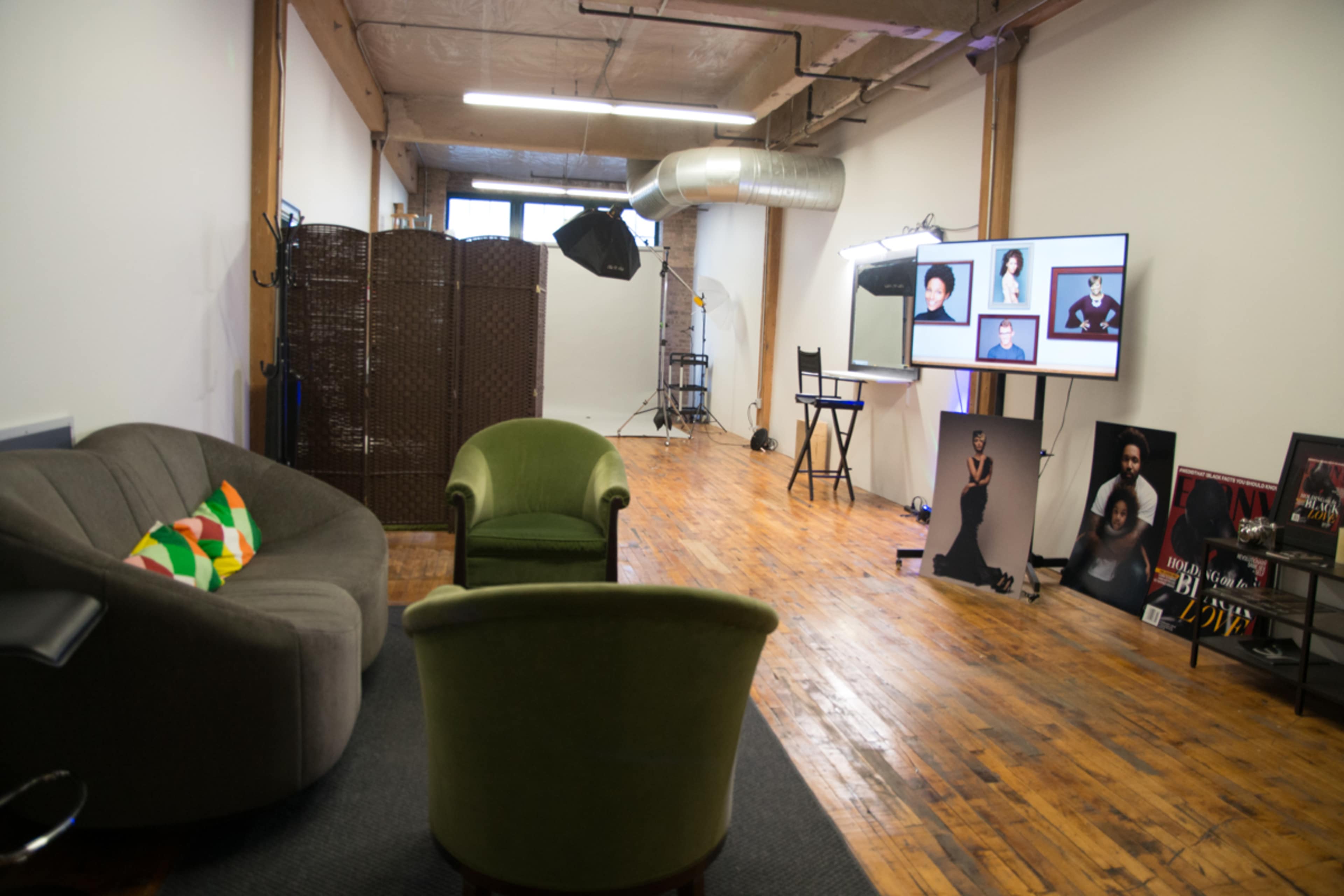 A spacious studio with wooden flooring, featuring a gray sofa, two green chairs, a television displaying portrait photos, and various lighting equipment set up for a photoshoot.