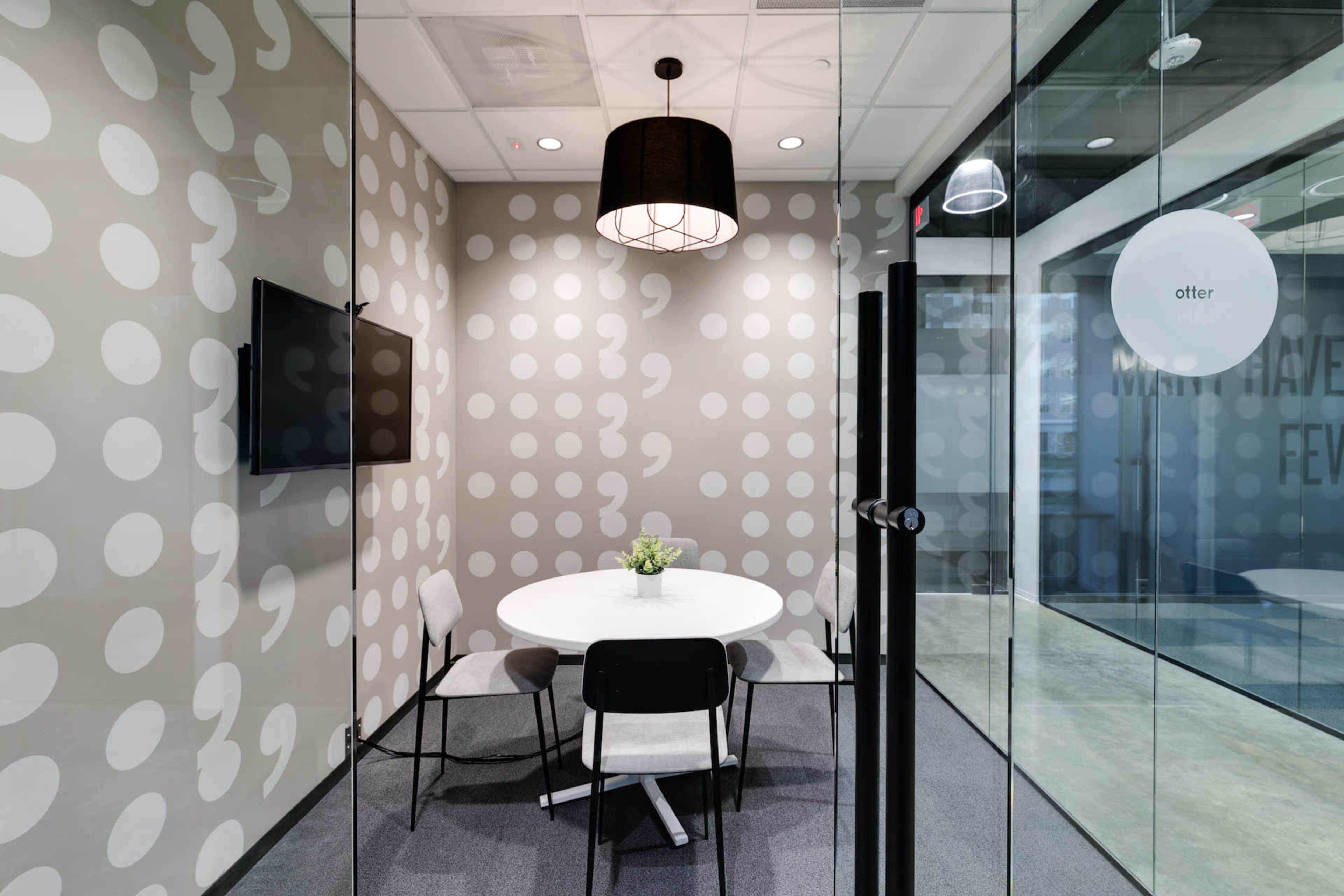 A glass-walled meeting room features a circular table surrounded by four chairs, with a patterned wall and a television mounted above.