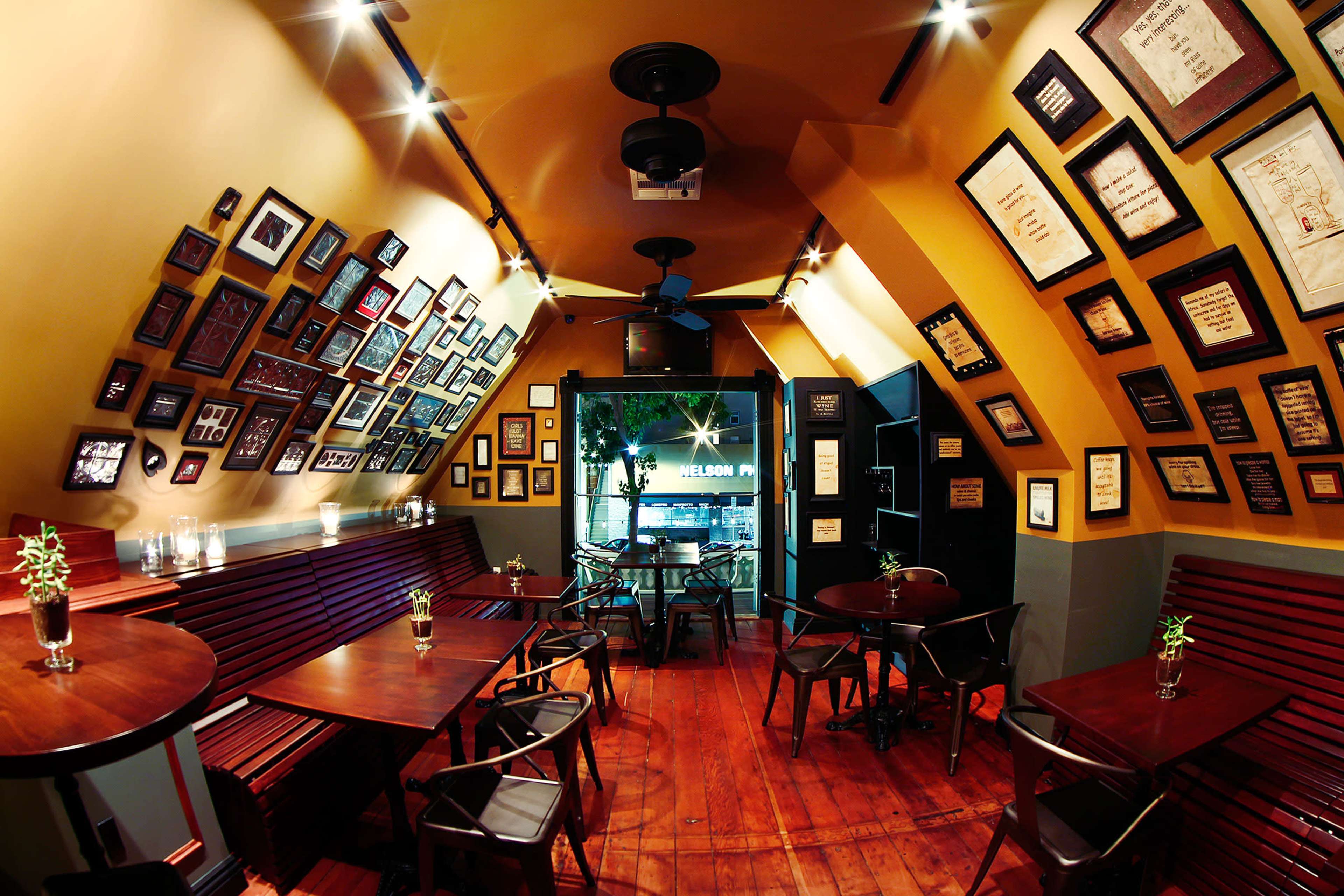 The image shows a small, cozy café with angled walls adorned with various framed pictures and certificates, featuring wooden tables and chairs arranged neatly.