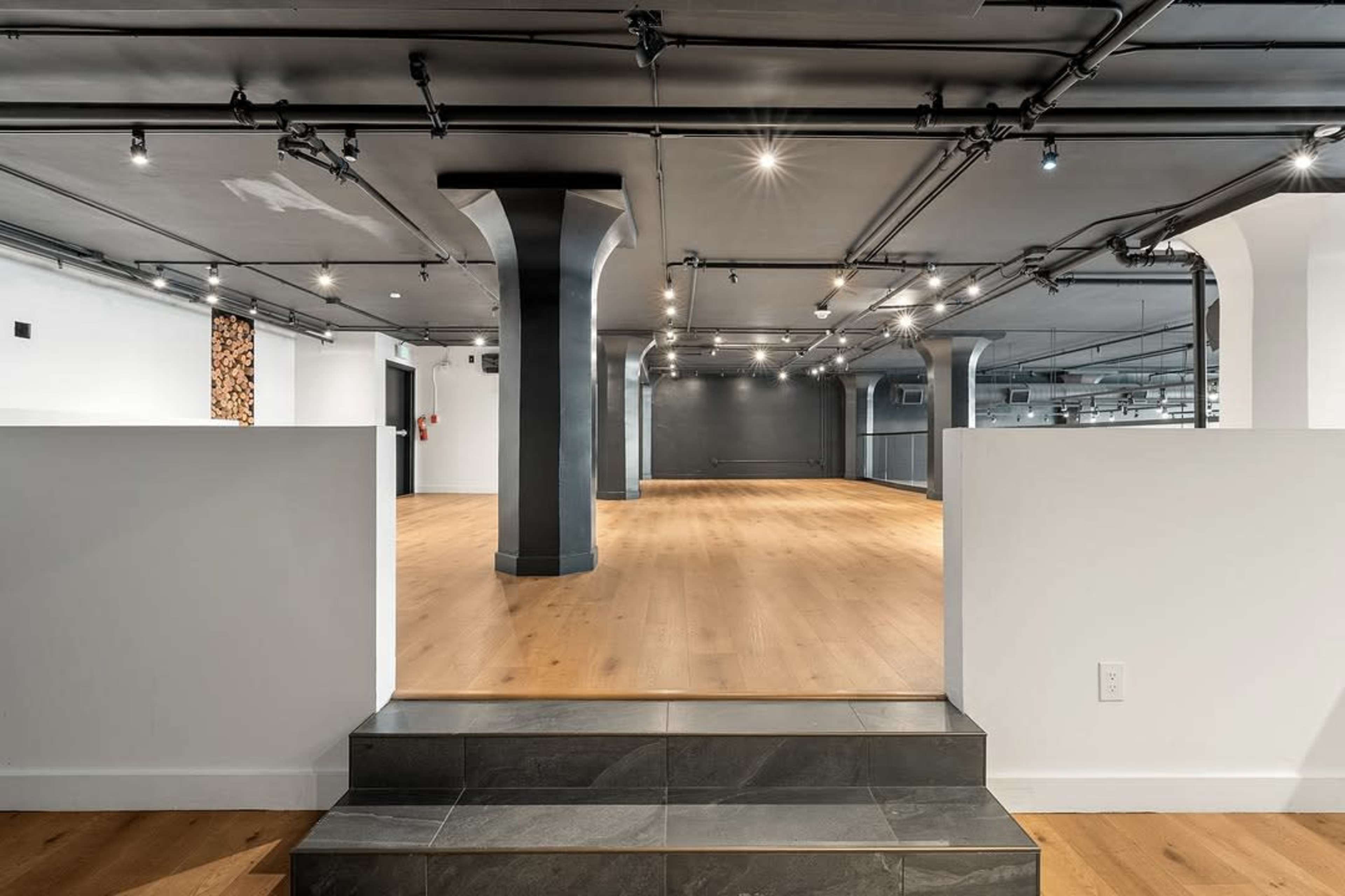 The image shows a spacious, modern interior with wooden flooring, exposed pipes, and a few columns, leading into an open area illuminated by overhead lights.