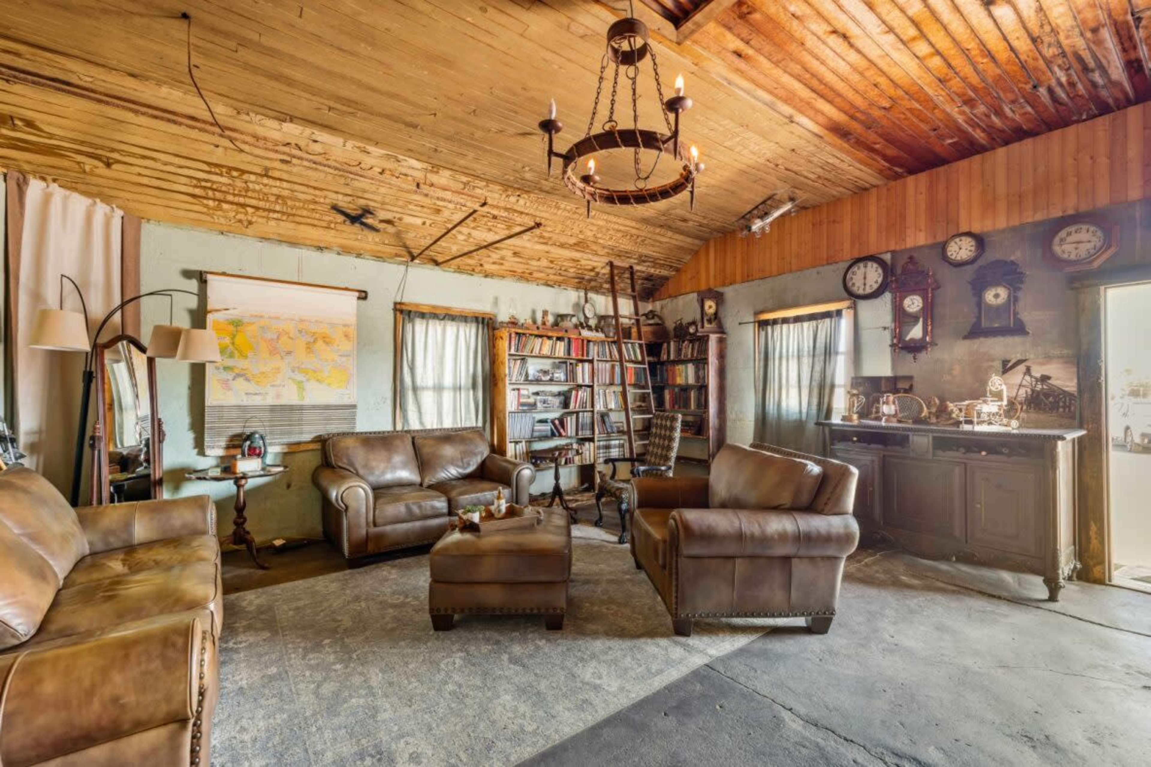 The image depicts a rustic living room featuring two leather sofas, a coffee table, a bookshelf, multiple wall clocks, and a small kitchenette.
