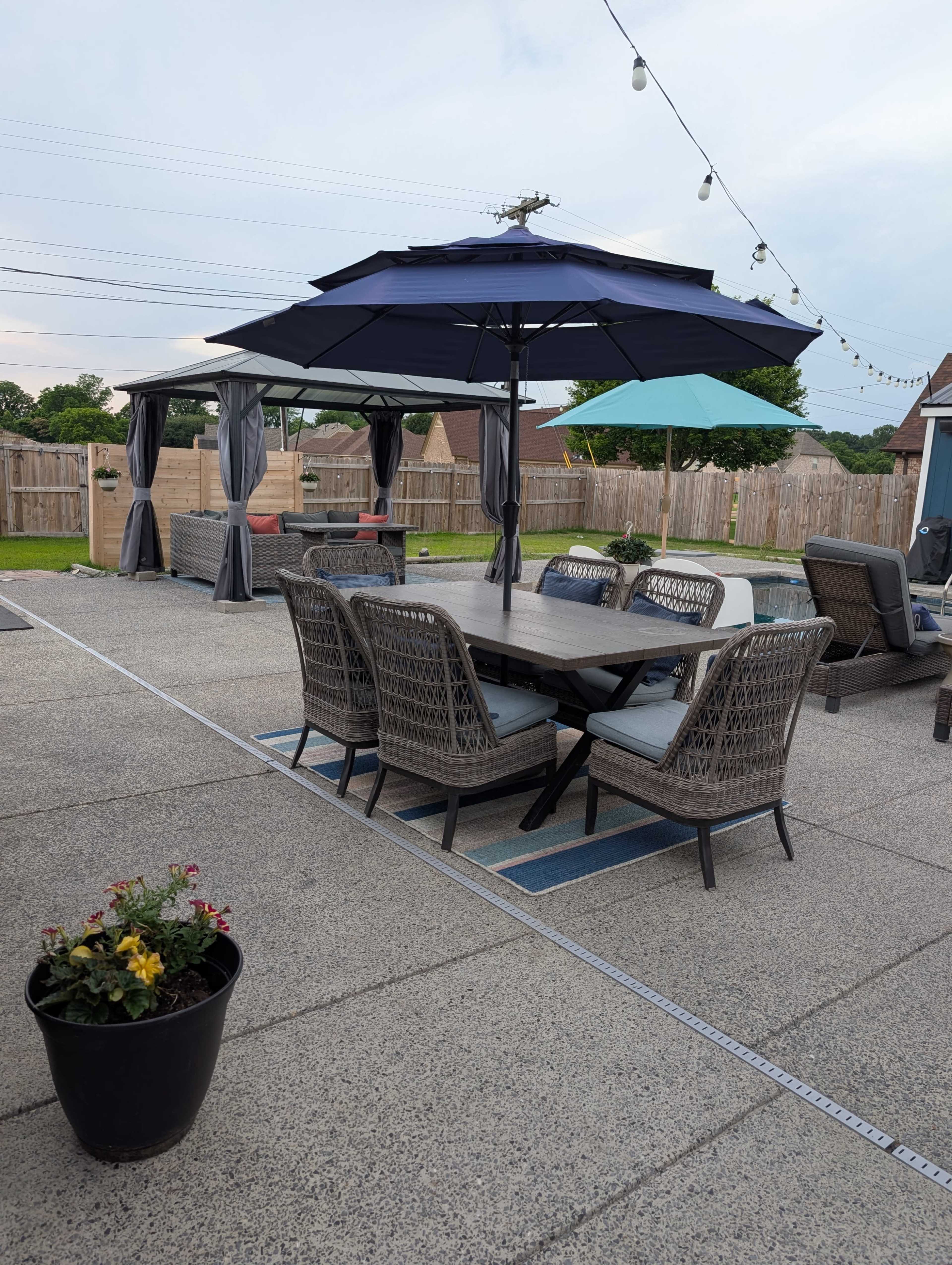 The image shows a patio area with a table and chairs covered by large umbrellas, surrounded by a fenced yard and a few potted plants.