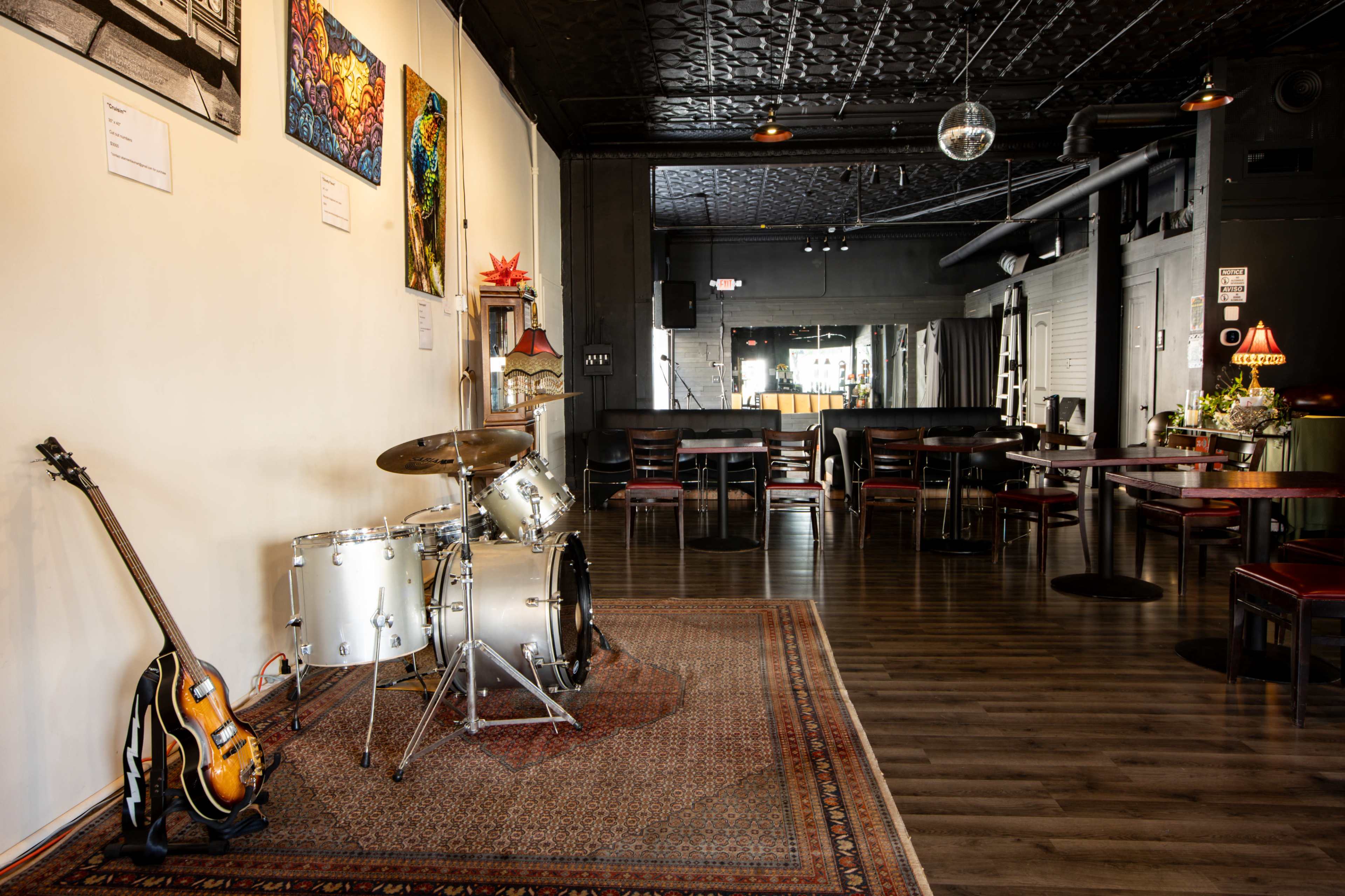 The image shows a music venue featuring a drum set and guitar on a rug near the entrance, with tables and chairs arranged throughout the space.
