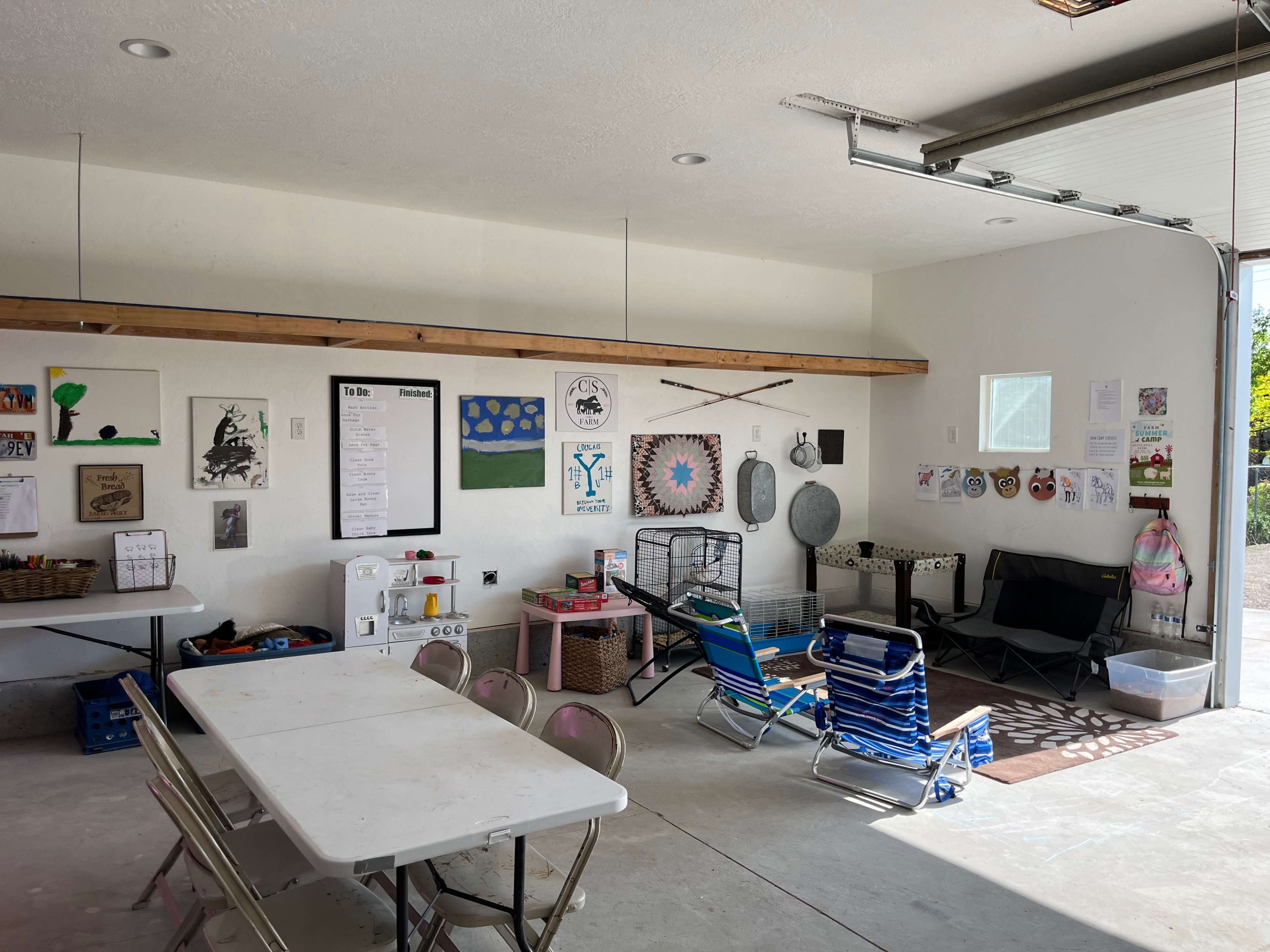 A spacious garage with white walls, a concrete floor, various artworks and notices hung on the walls, and a table with chairs, along with seating and storage items scattered around.