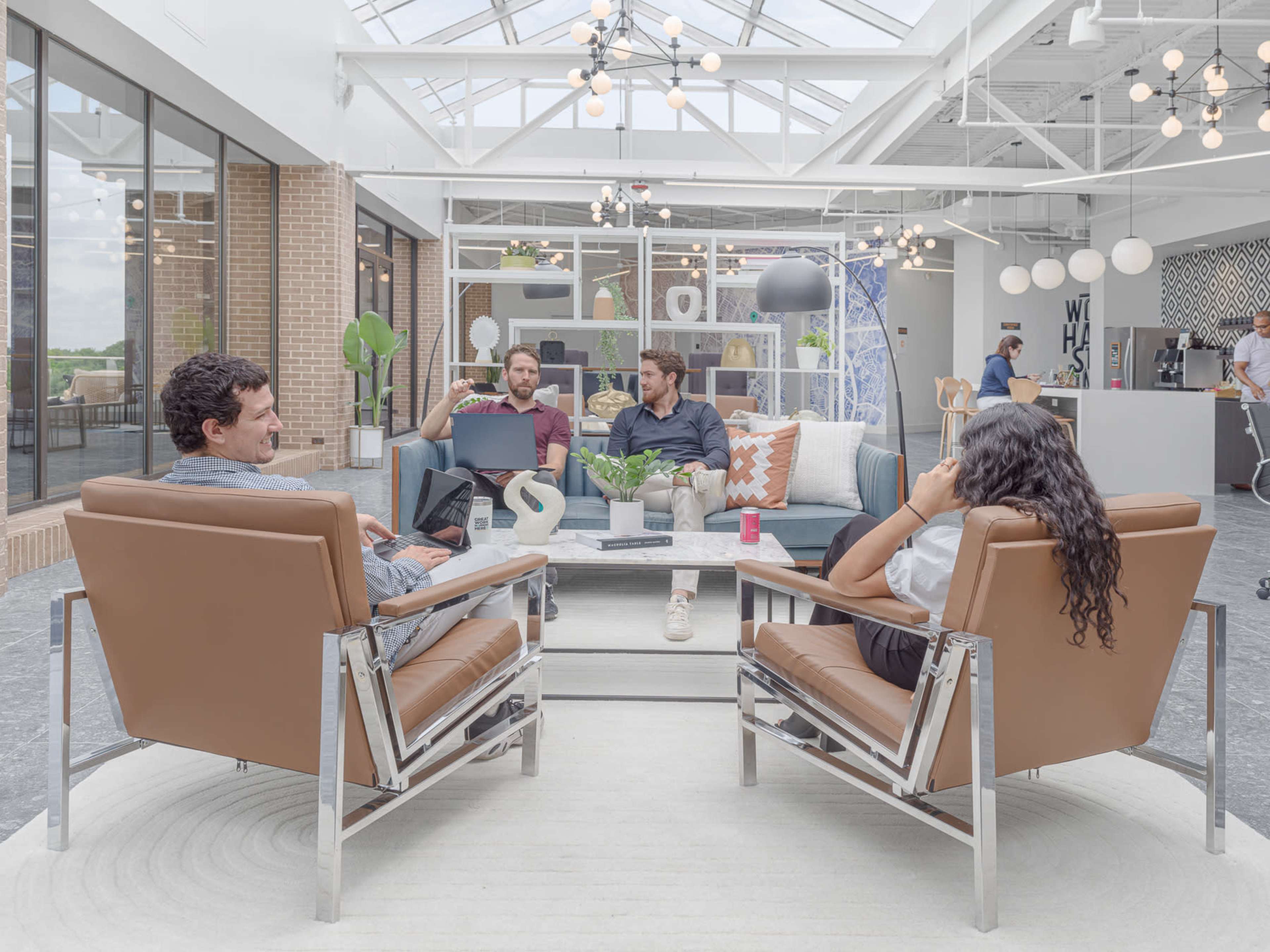 A modern co-working space features four people engaged in discussion, seated in leather chairs around a central seating area with a coffee table.