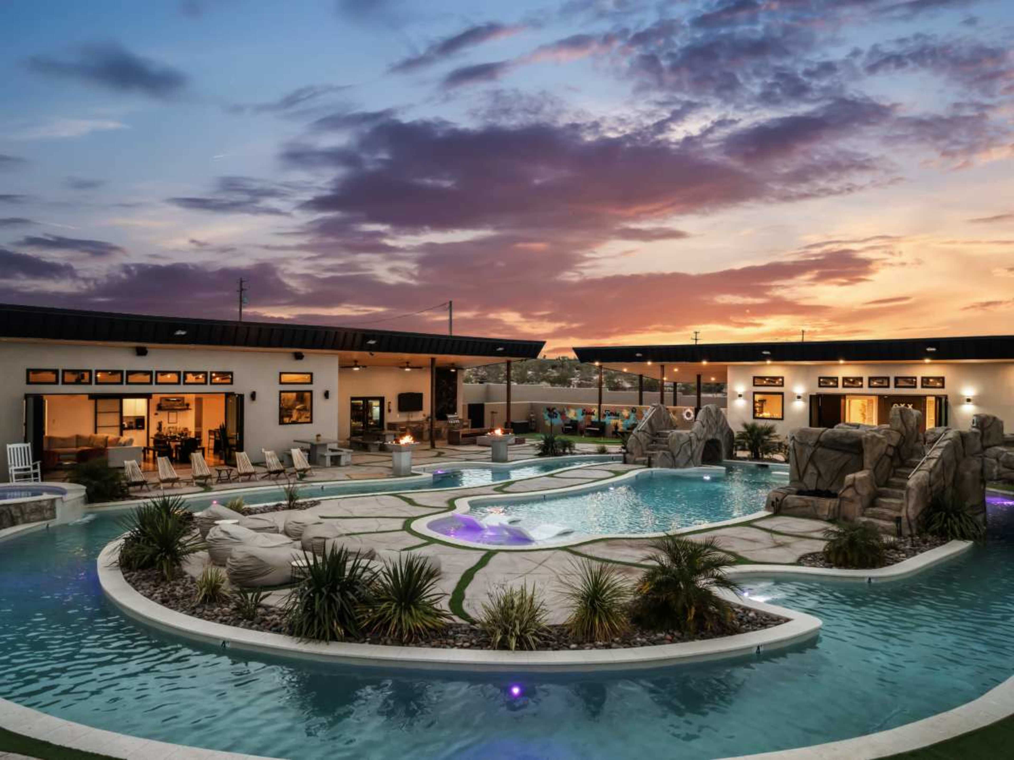 The image shows a spacious backyard pool area with rock formations and landscaped greenery, set against a vibrant sunset sky.