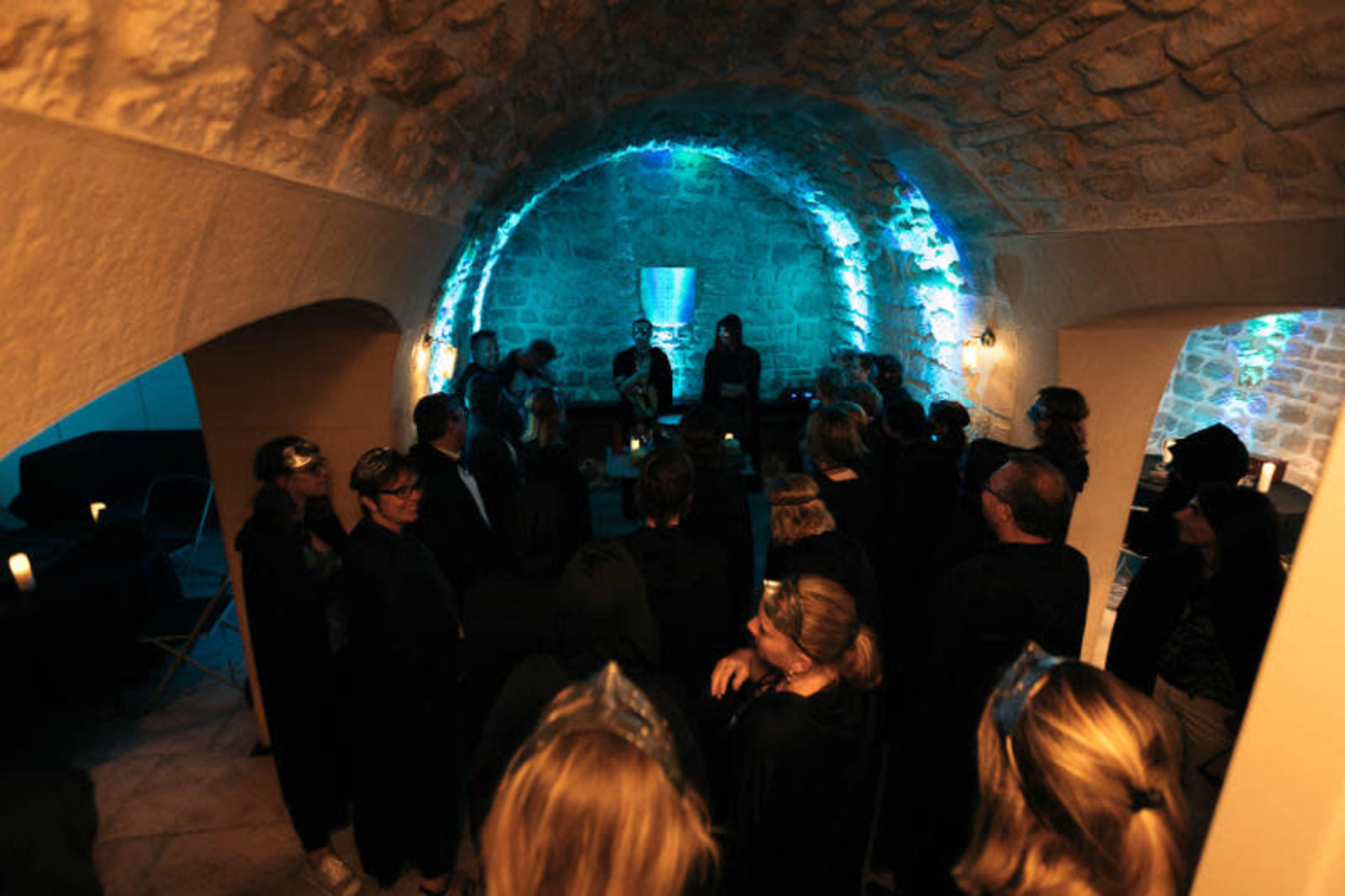 A group of people in dark clothing is gathered in a dimly lit stone-arched room with blue lighting, listening to musicians in the background.