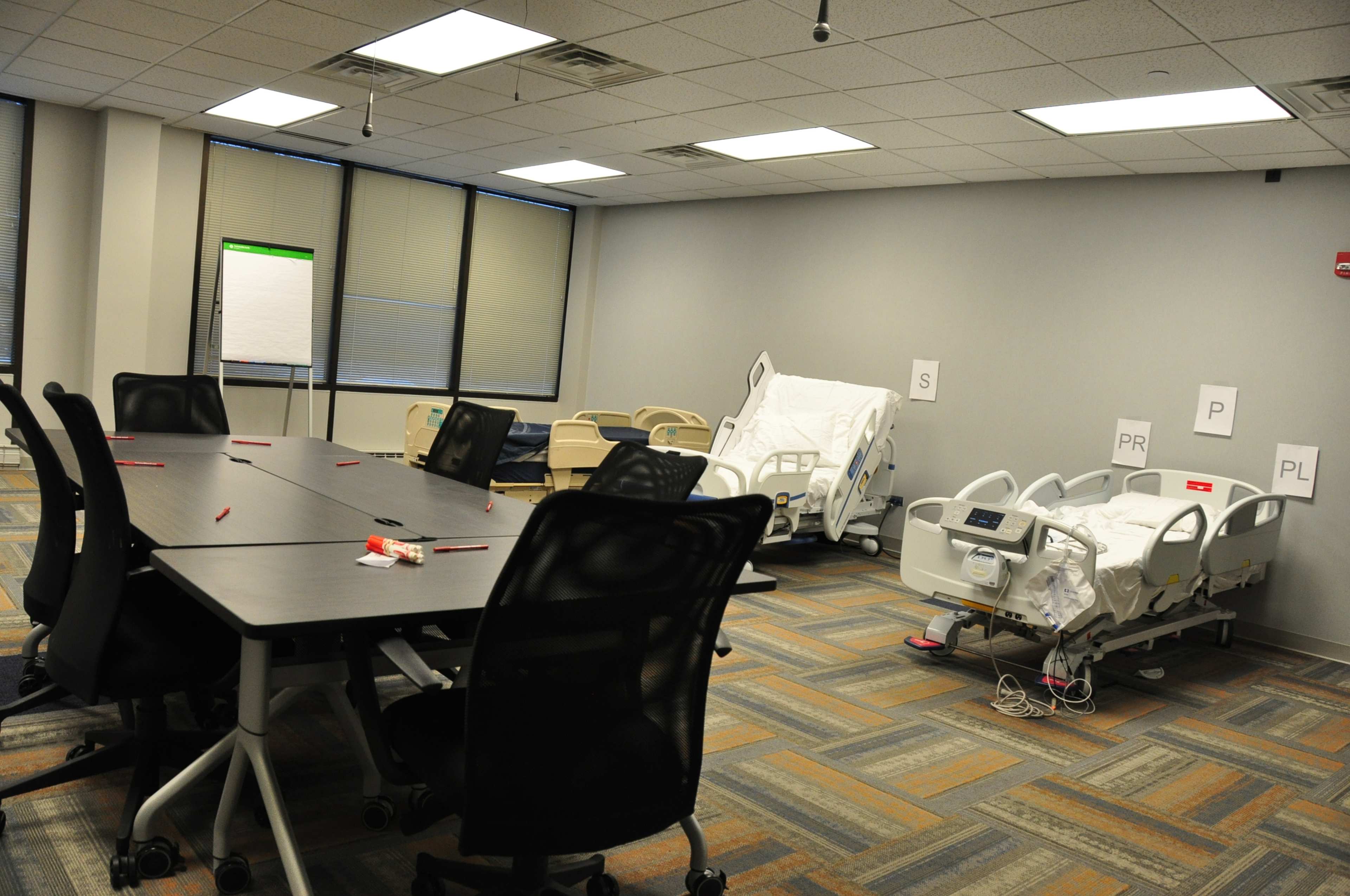 The image shows a medical training room with a large table, several chairs, and two hospital beds labeled with letters.