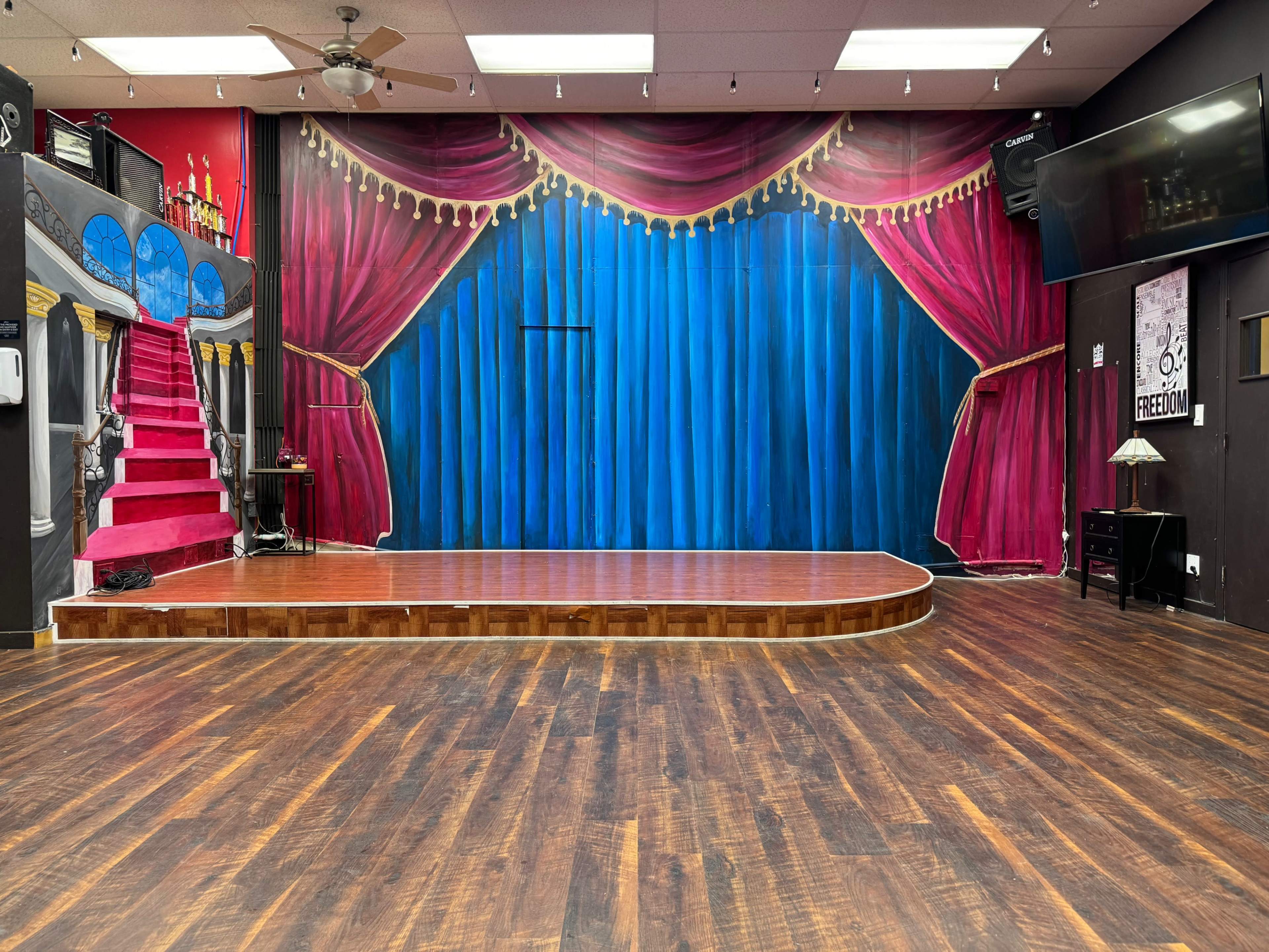 A stage with a blue curtain backdrop and red drapery, surrounded by a wooden floor and a staircase on the left.
