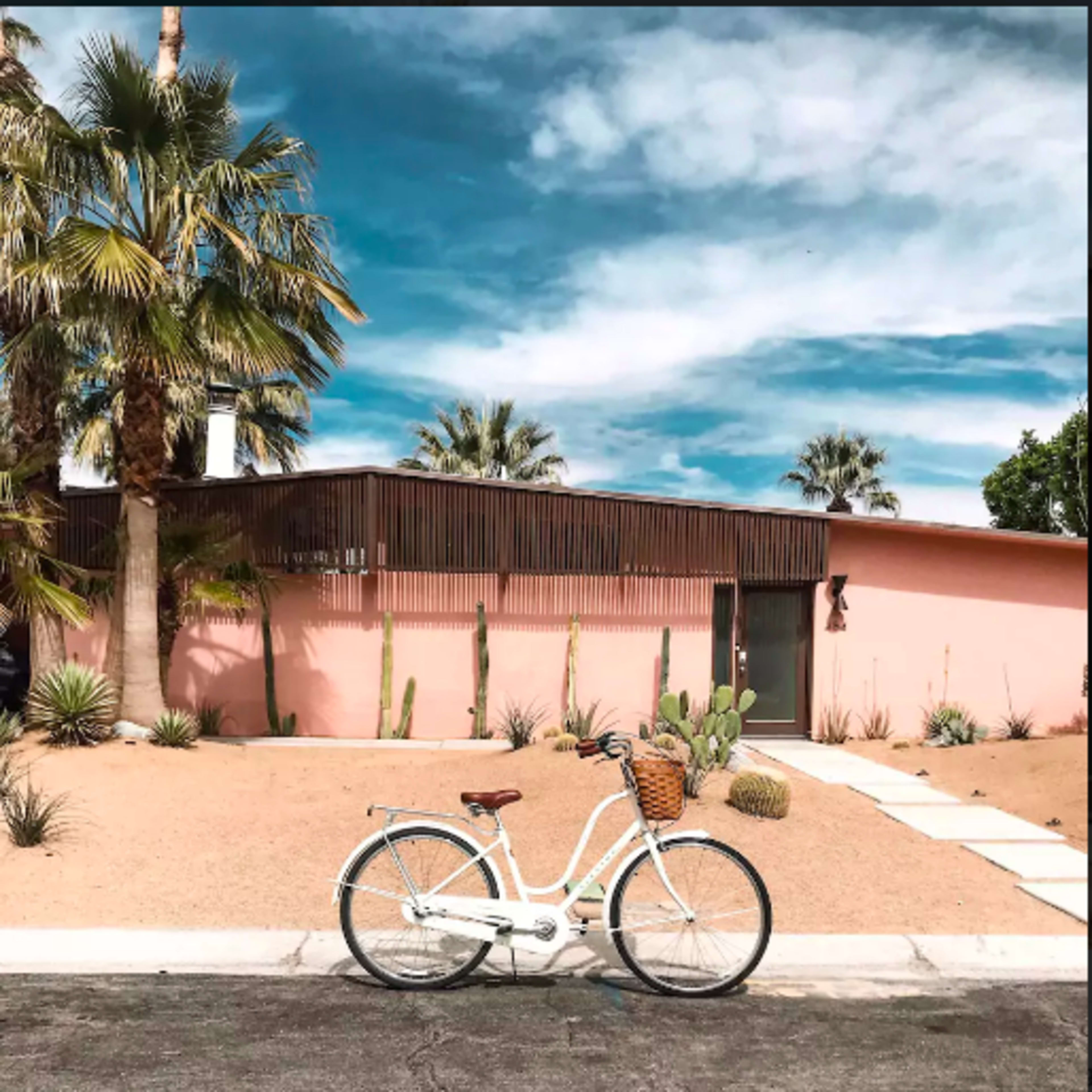 A white bicycle is parked next to a pink house surrounded by palm trees and desert landscaping.