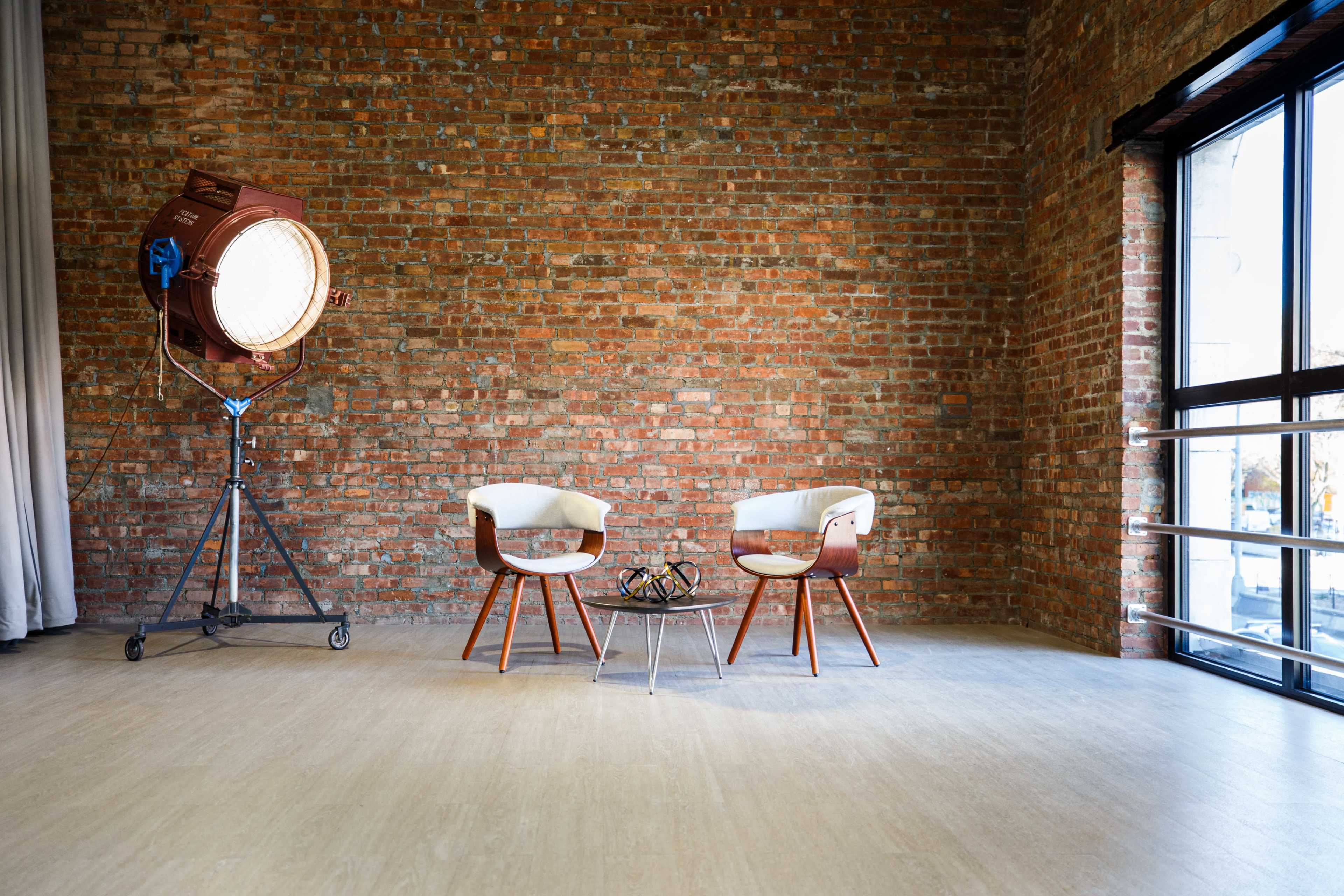 A minimalist interior featuring two white chairs with wooden legs, a glass table, and a large studio light against a brick wall.