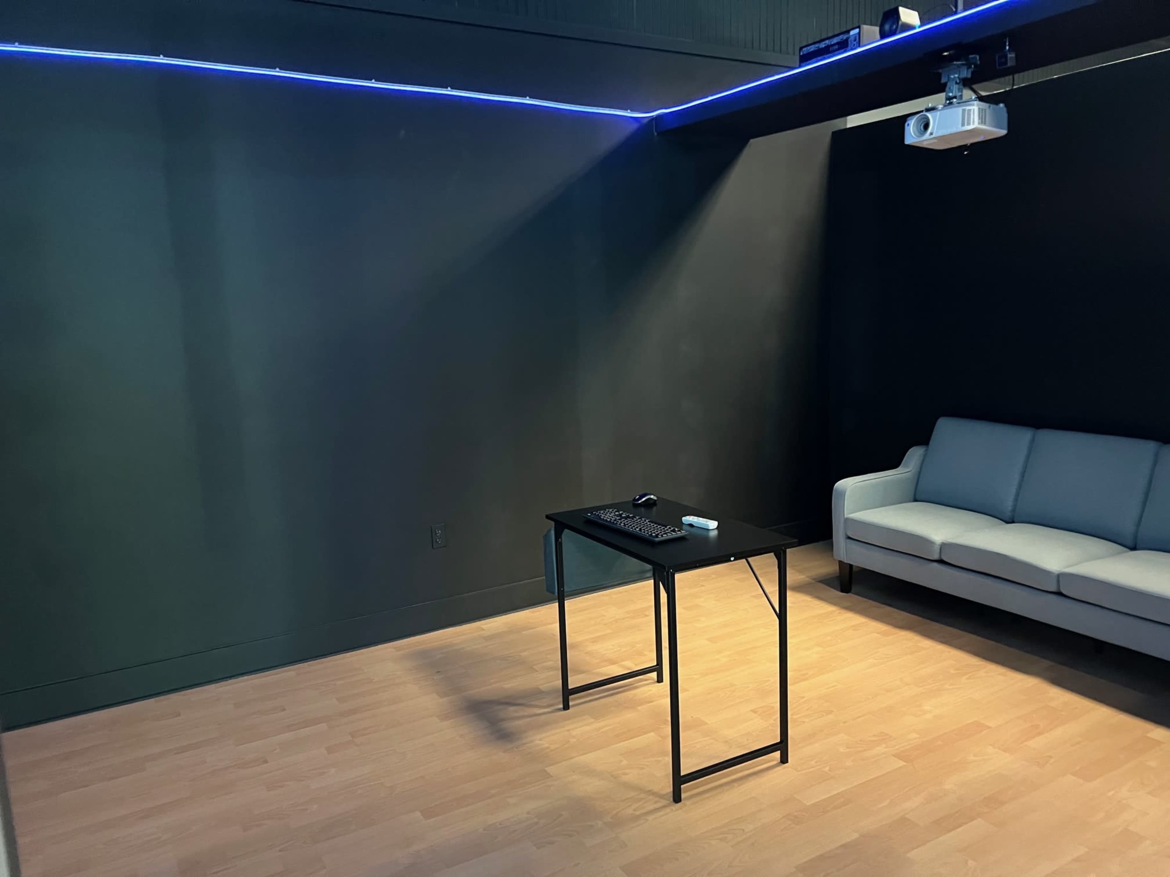 The image shows a dimly lit room with a black wall, a projector mounted on the ceiling, a small desk with a keyboard and remote, and a gray sofa.