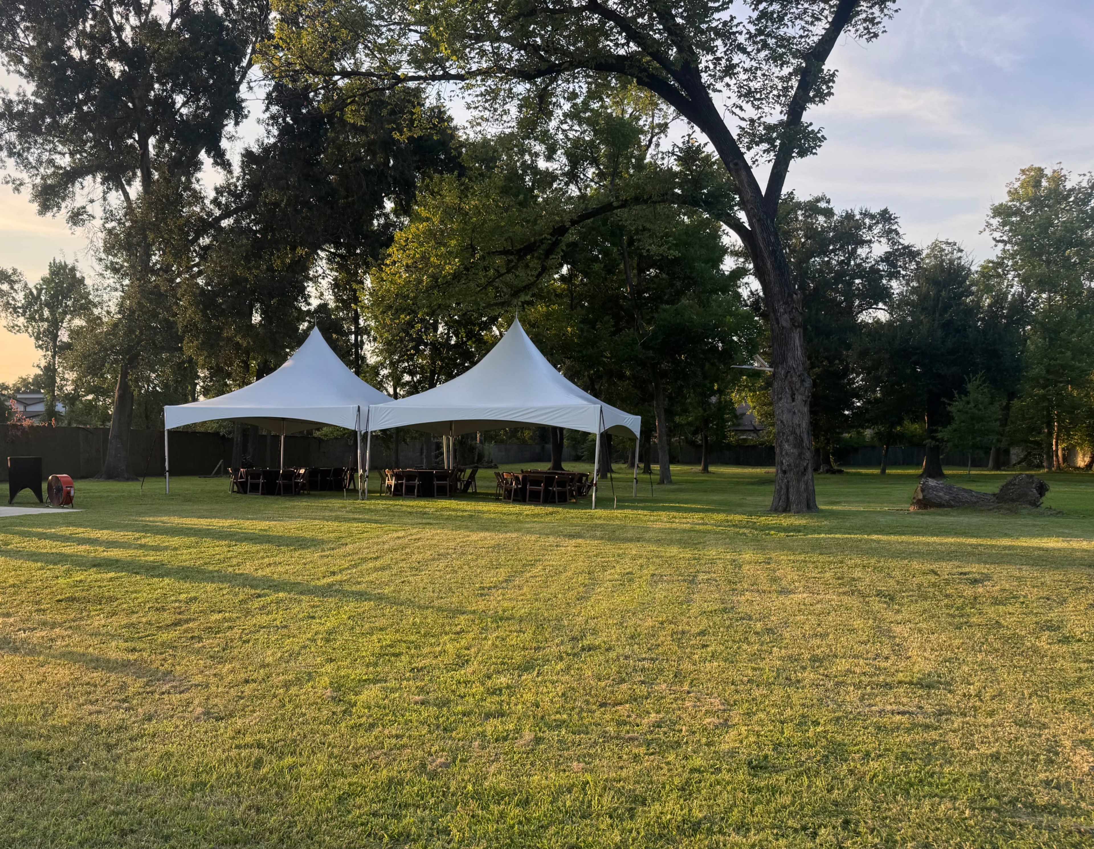 A large white tent is set up in a grassy area surrounded by trees, with tables and chairs arranged underneath.