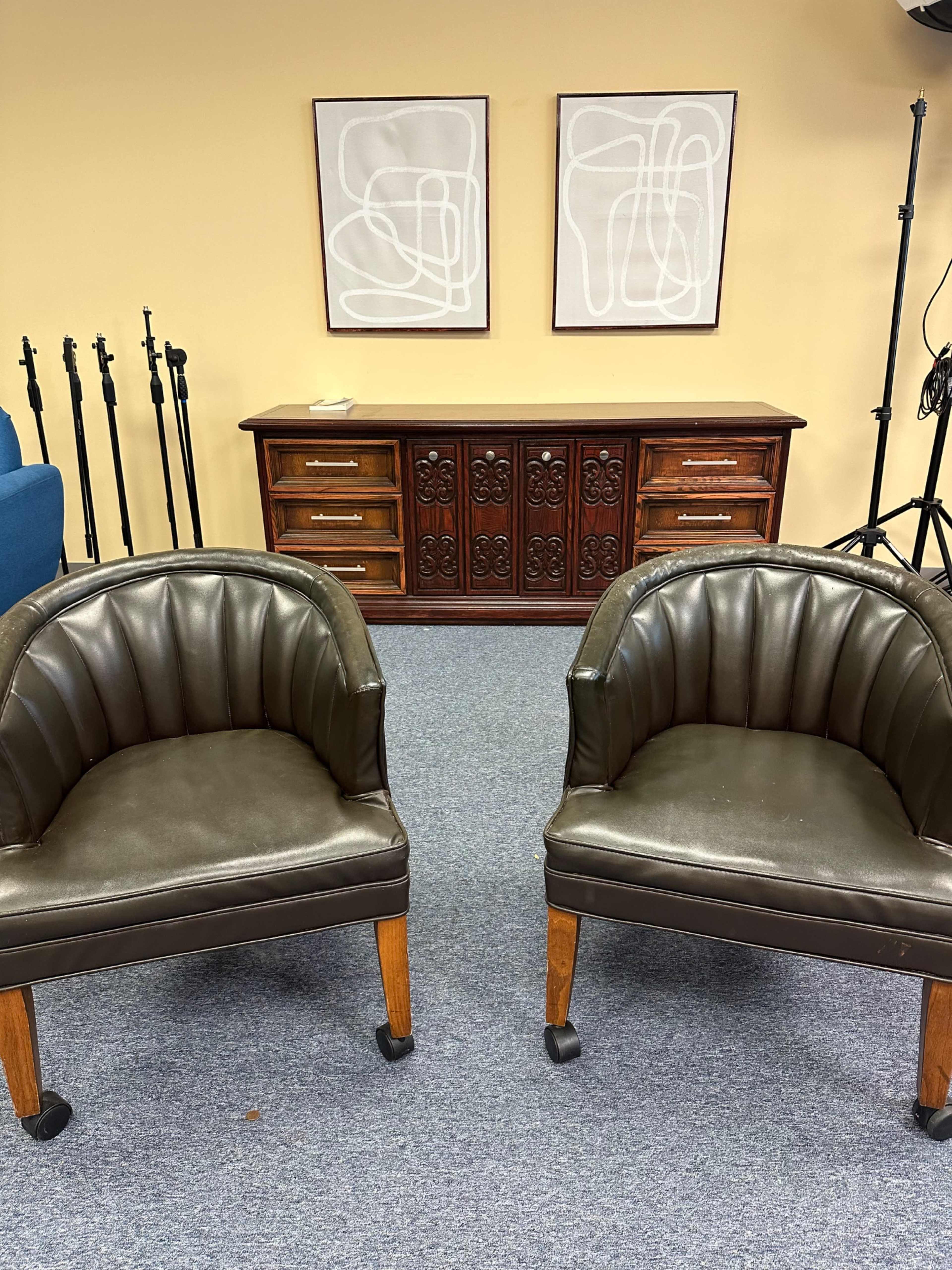 Two brown upholstered chairs are positioned in front of a wooden cabinet with ornate designs, flanked by two framed abstract art pieces on a beige wall.