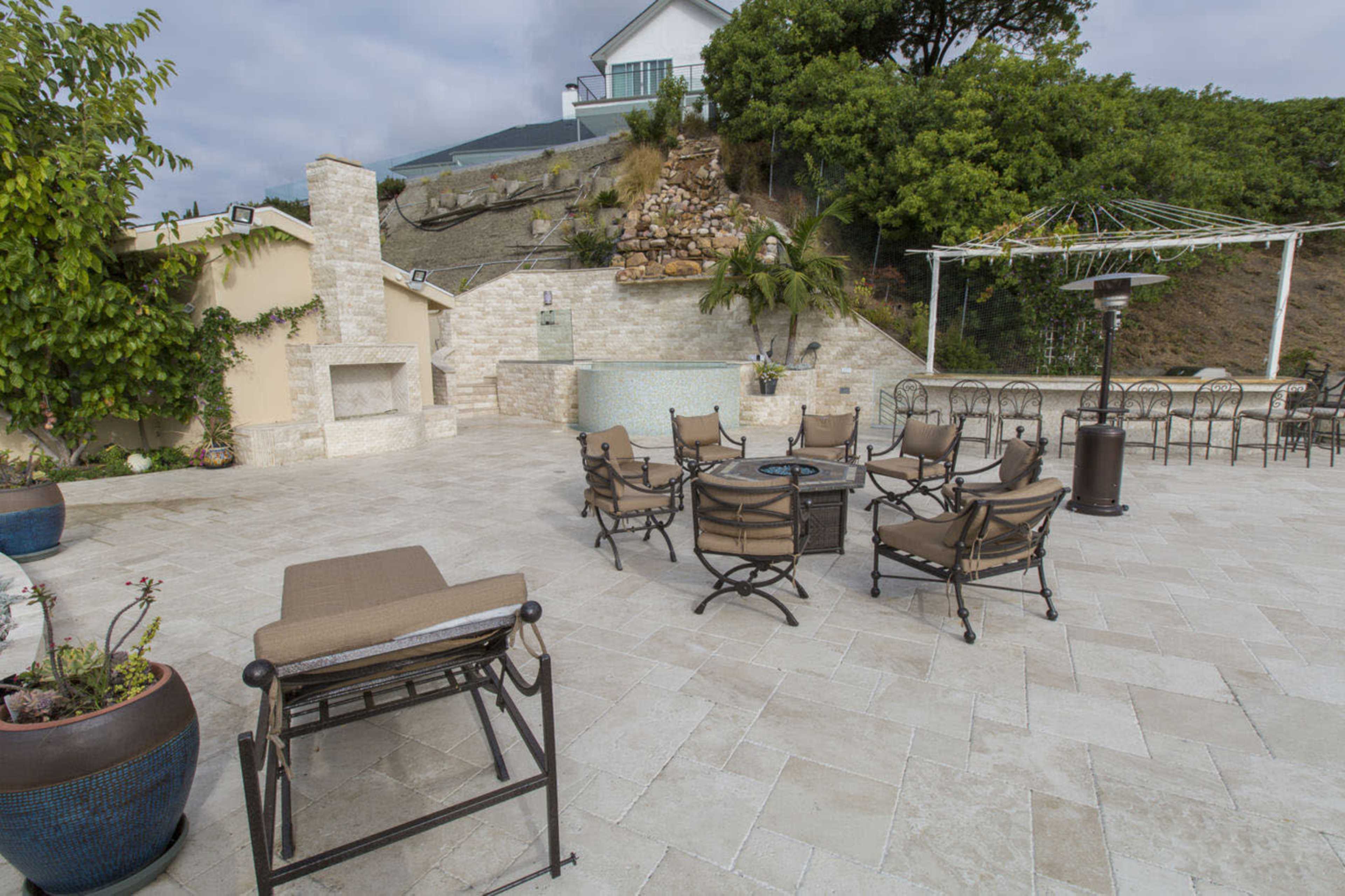 The image shows a spacious outdoor patio area with a seating arrangement around a fire pit, bordered by landscaping and an outdoor kitchen.