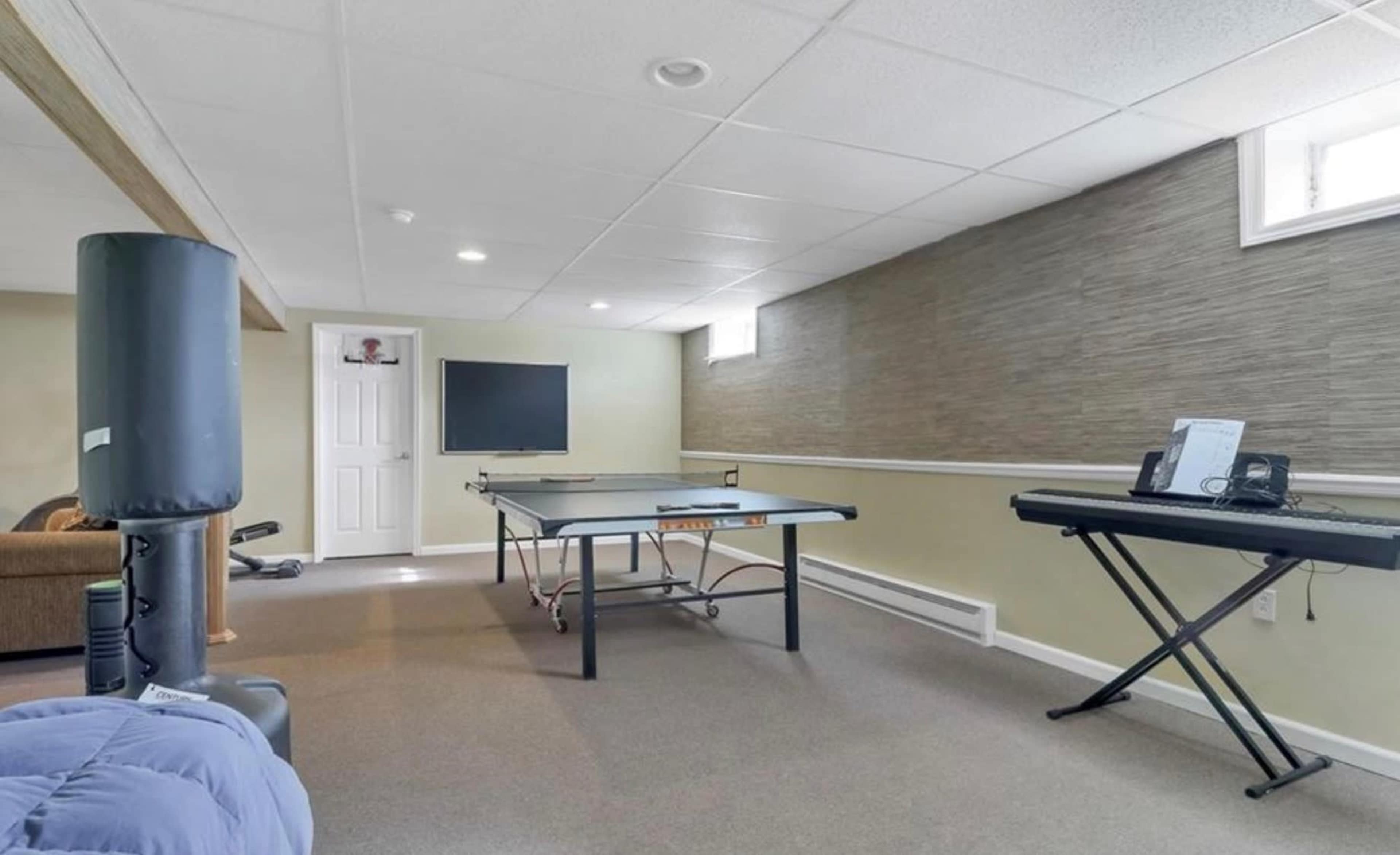 A basement room featuring a ping pong table, a keyboard on a stand, a television mounted on the wall, and a door leading to another area.