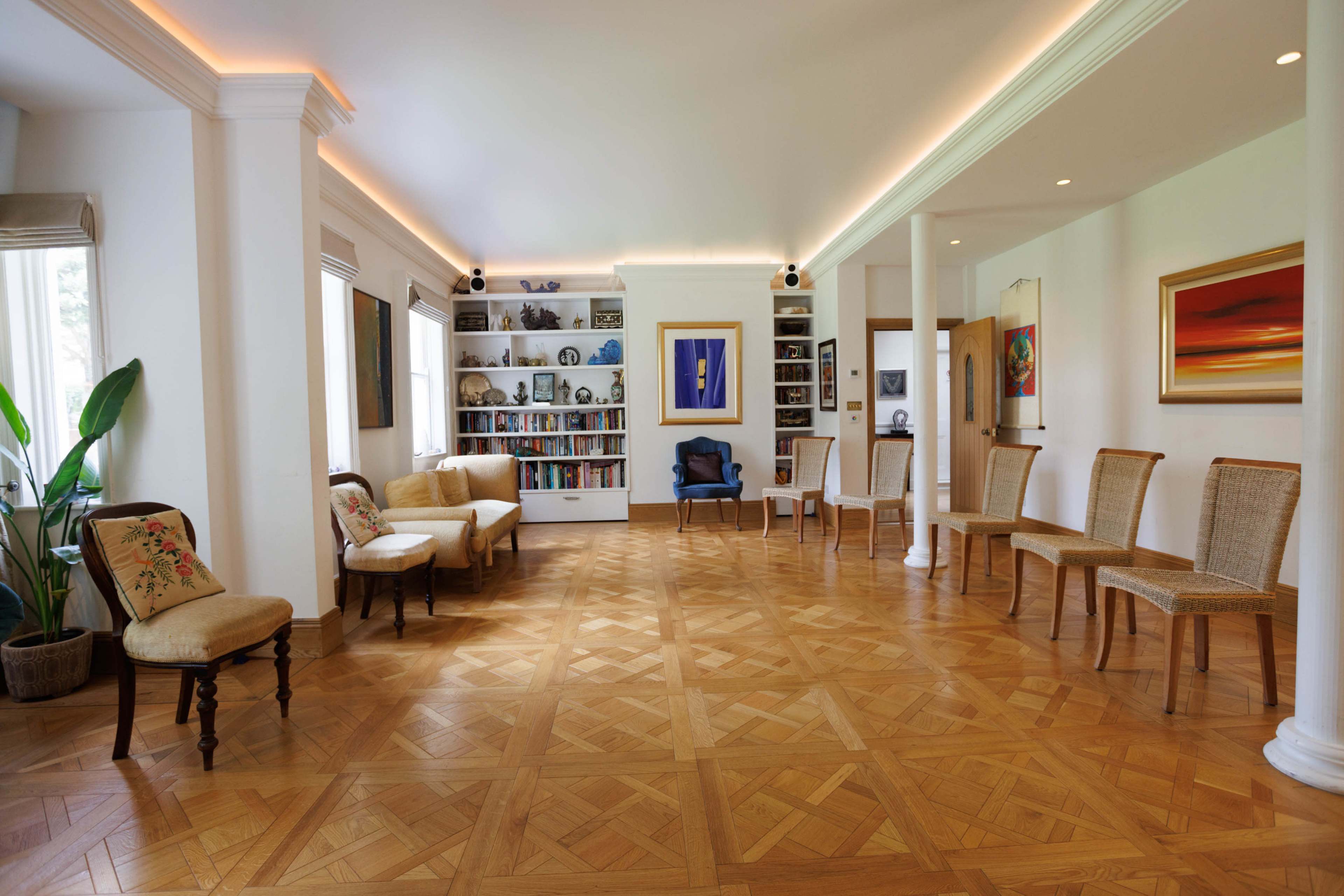 A spacious room with wooden flooring, featuring a seating area with upholstered chairs and a bookshelf displaying various items along the walls.