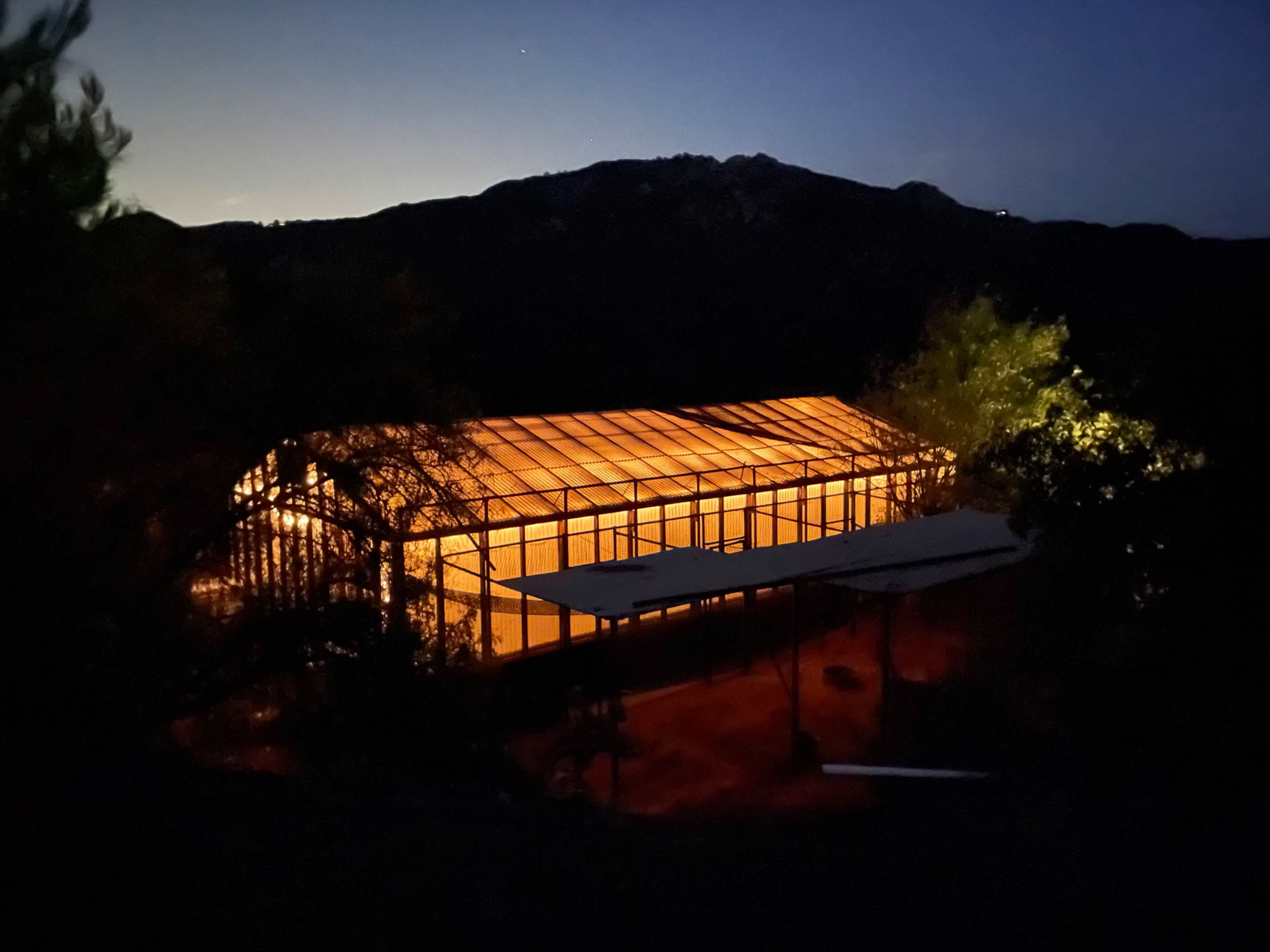 A greenhouse illuminated from within stands against a night backdrop of mountains.