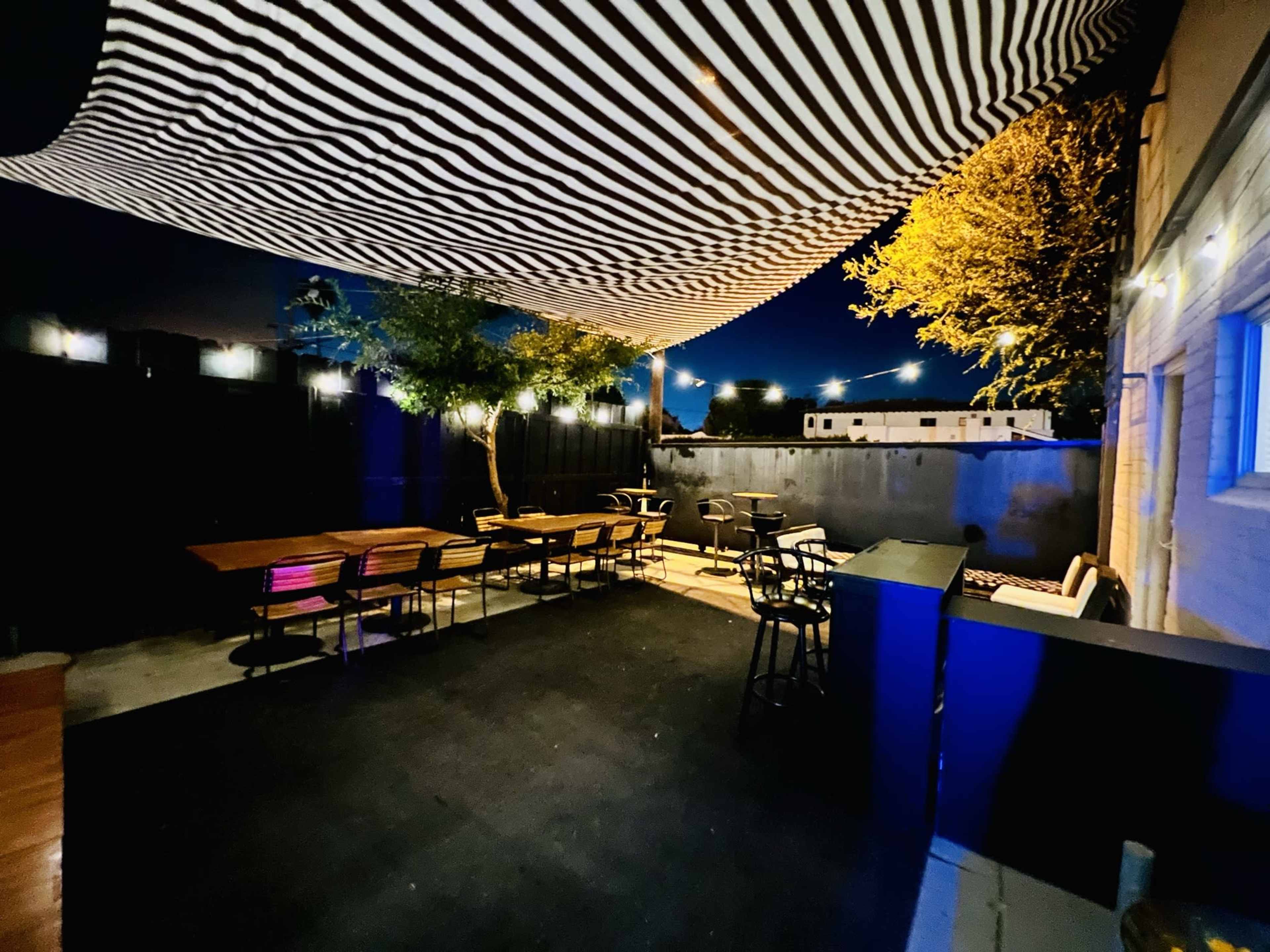 An outdoor patio area is illuminated at night, featuring striped canopies, wooden tables, and chairs, with multiple light sources creating a cozy ambiance.