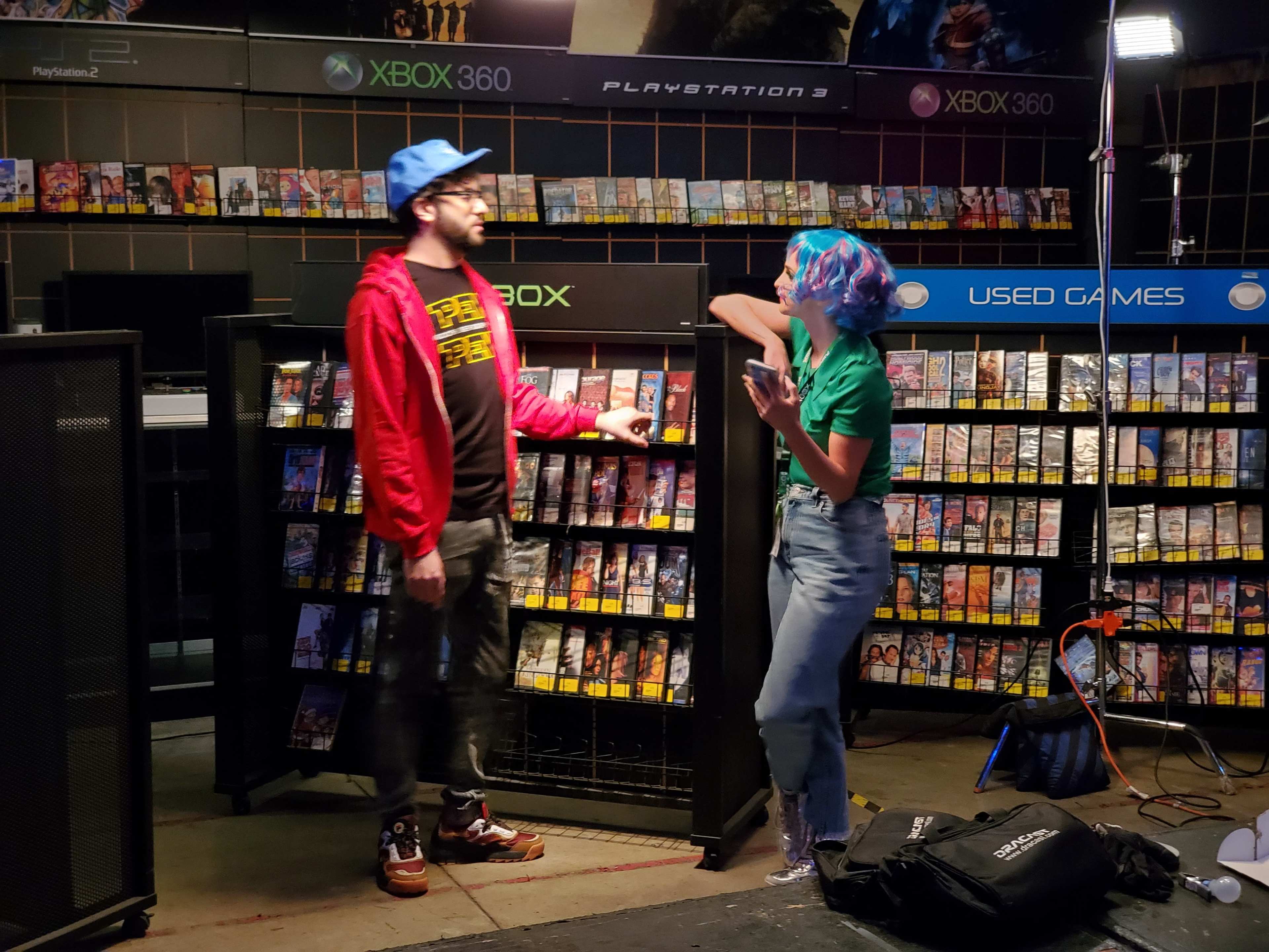 A man in a red jacket and blue cap talks to a woman with blue hair wearing a green shirt, standing near shelves filled with DVD cases for video games.