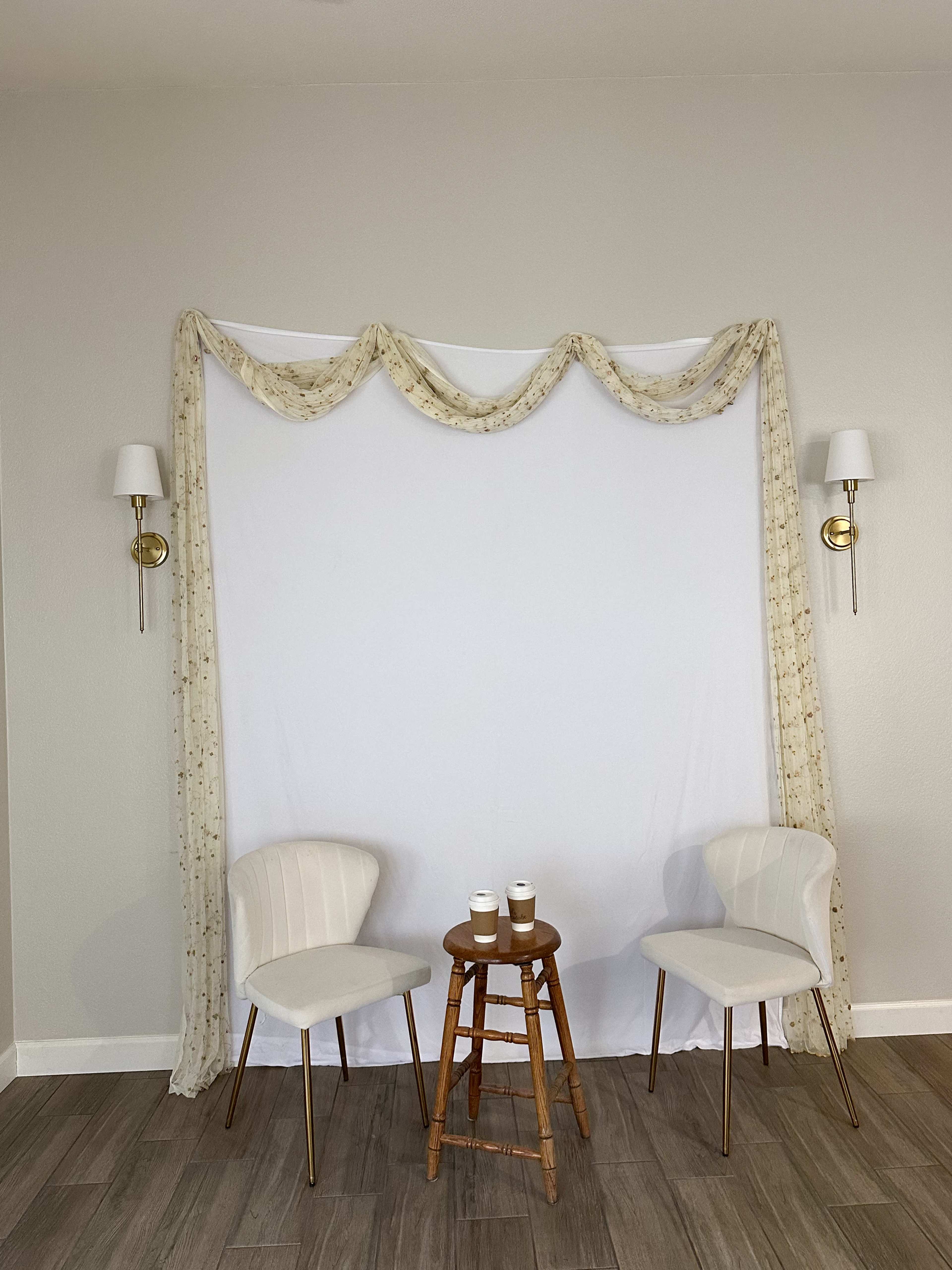 The image shows two white chairs on either side of a small wooden stool in front of a plain white backdrop draped with fabric and wall sconces.