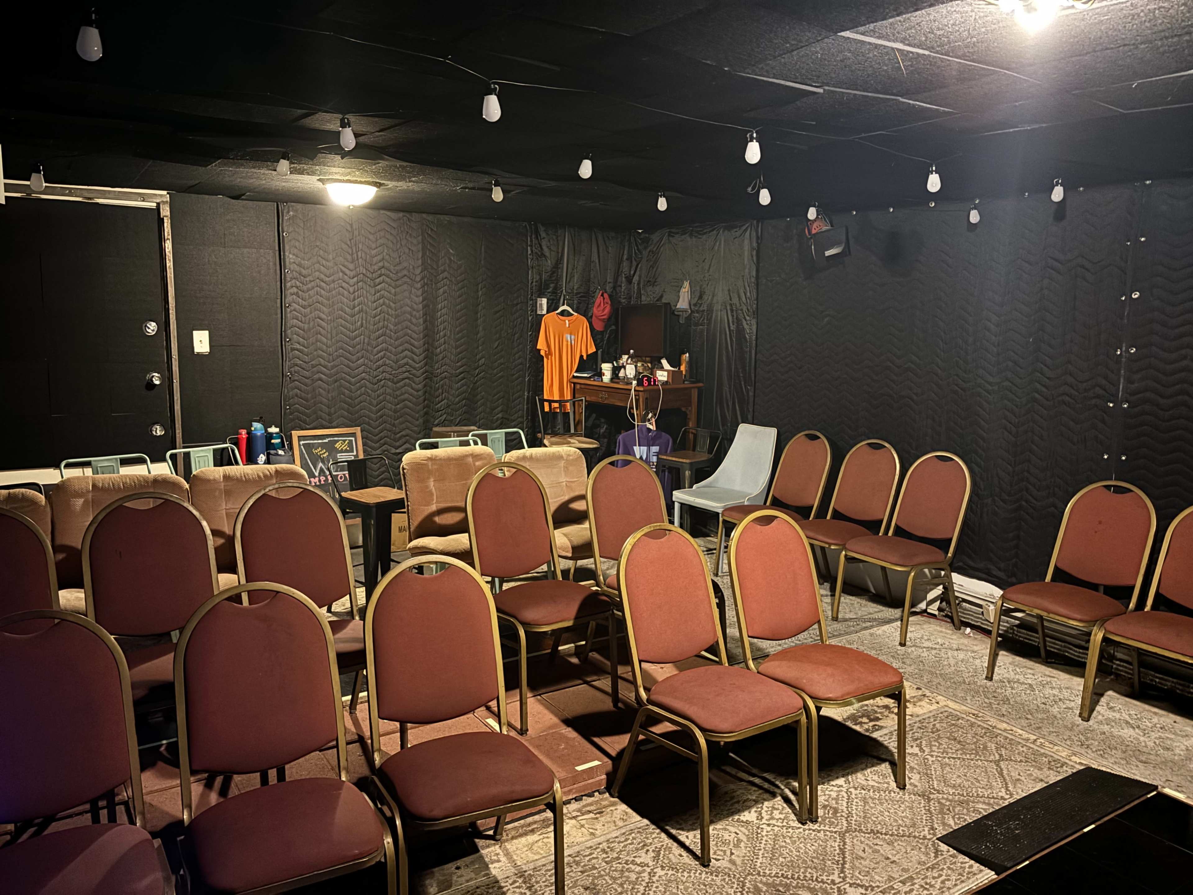 A small, dimly lit performance space with rows of red chairs facing a stage area adorned with equipment and props.