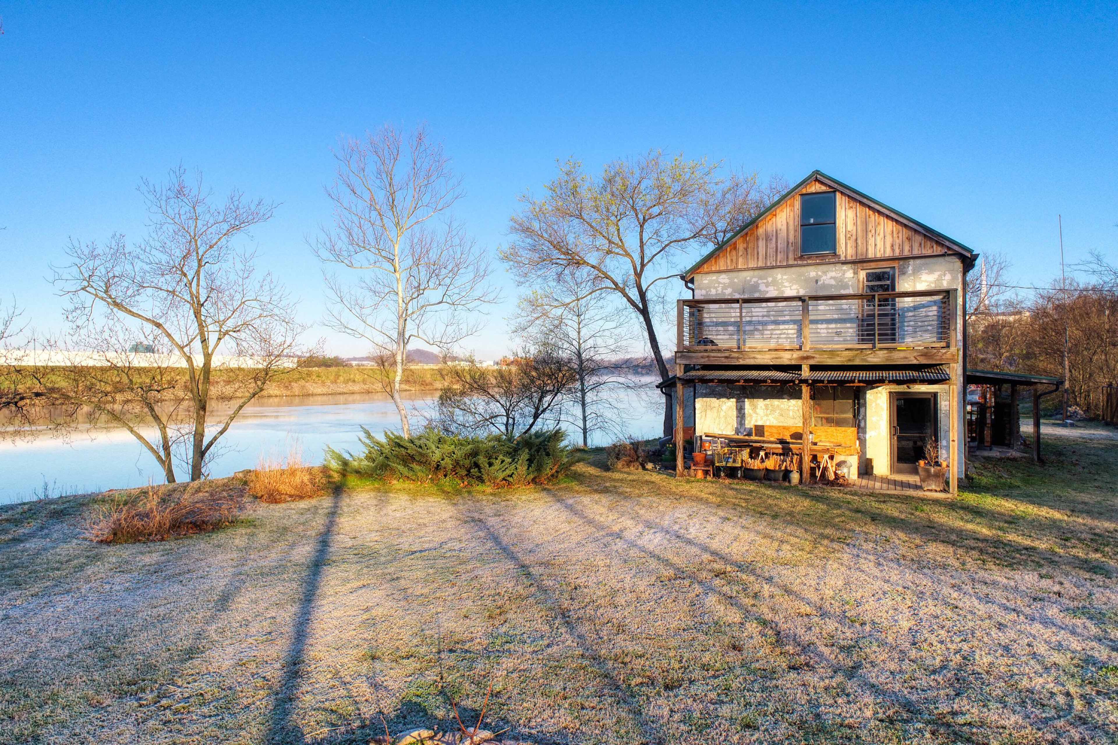 A two-story wooden house stands near a calm body of water, with sparse trees and a frost-covered lawn in the foreground.