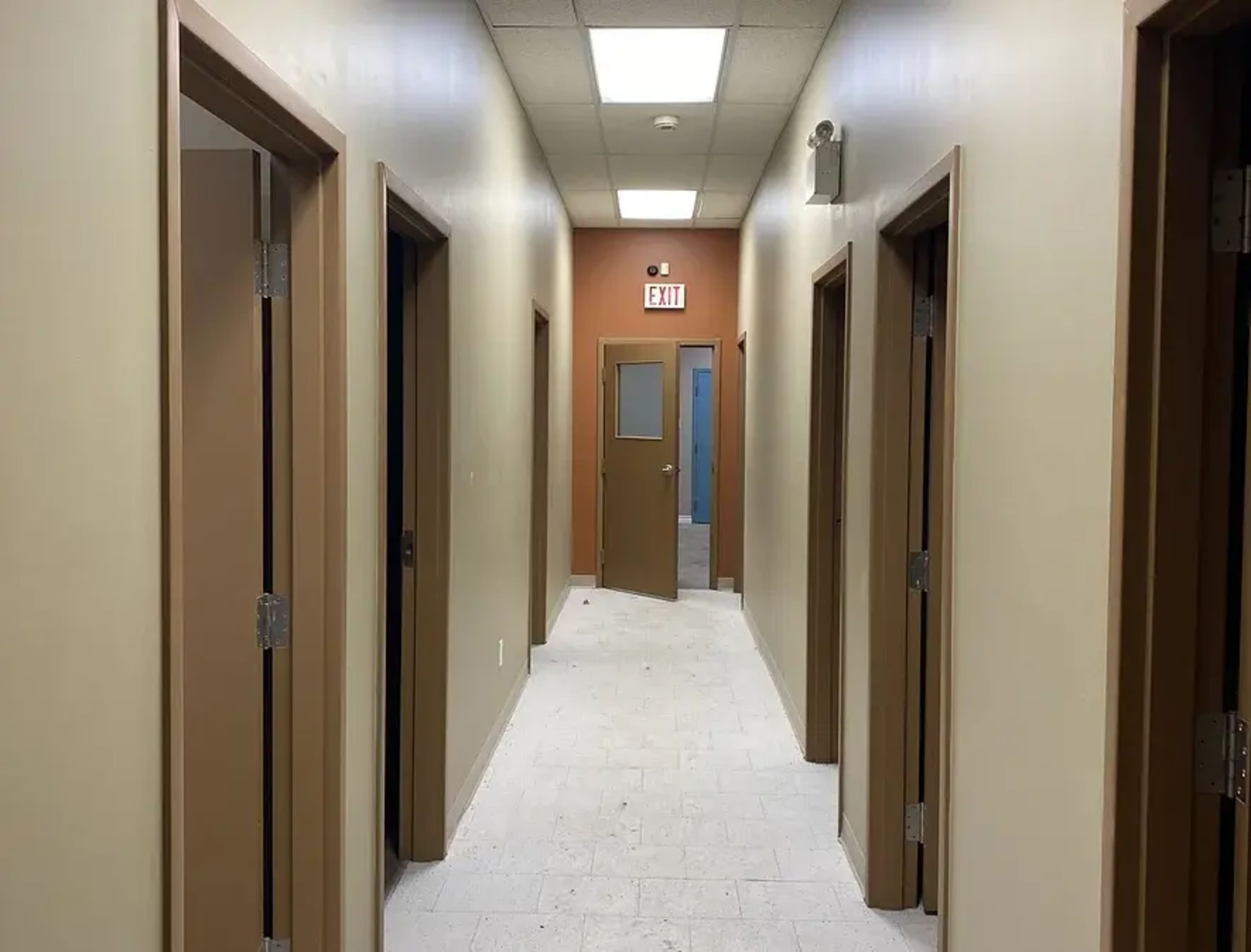 A long, narrow hallway with several doors on either side and a door at the end leading to an exit.
