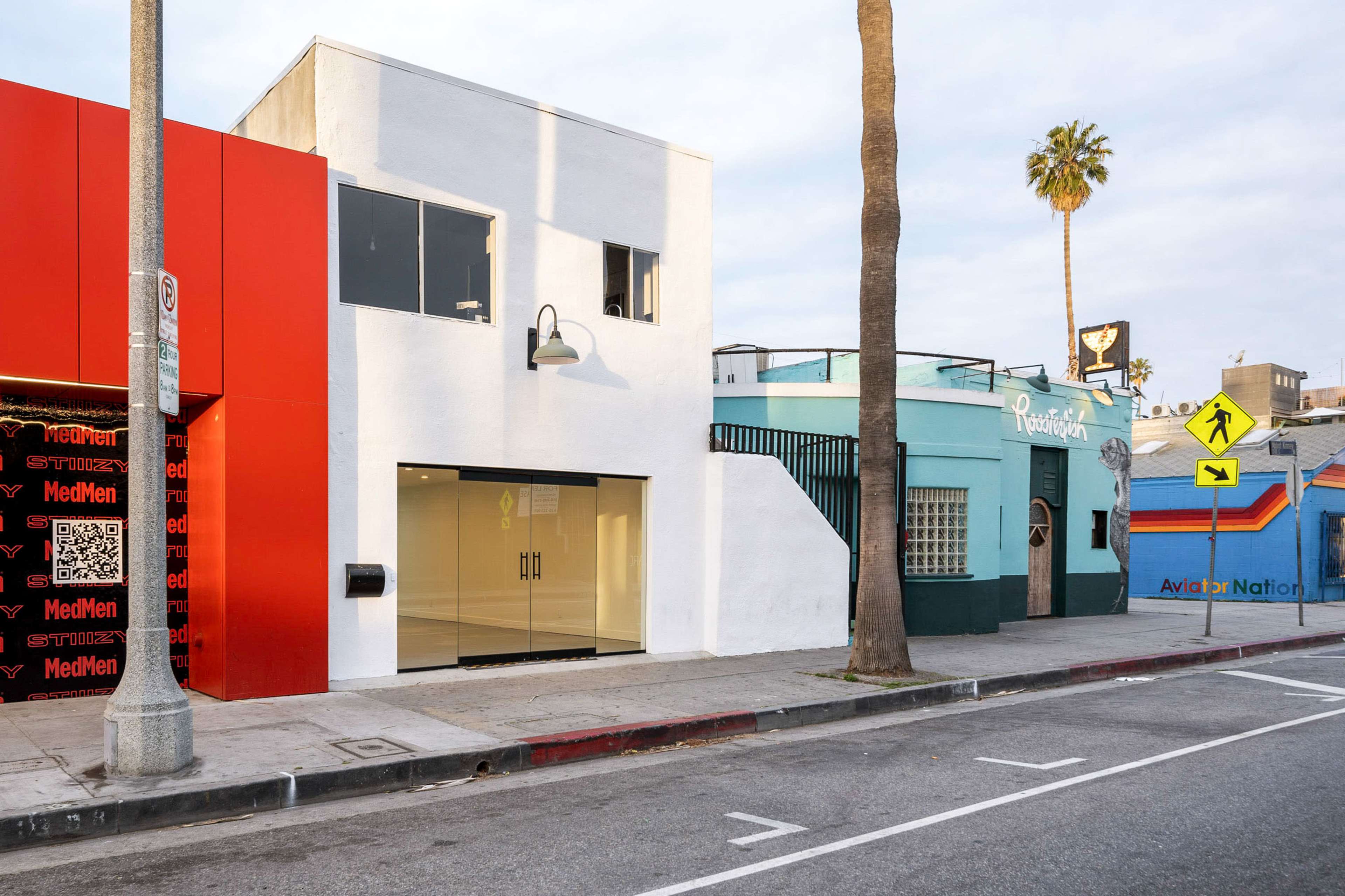 The image shows a street scene featuring a two-story white building next to colorful storefronts, including a red building and a teal structure, set under palm trees along a sidewalk.