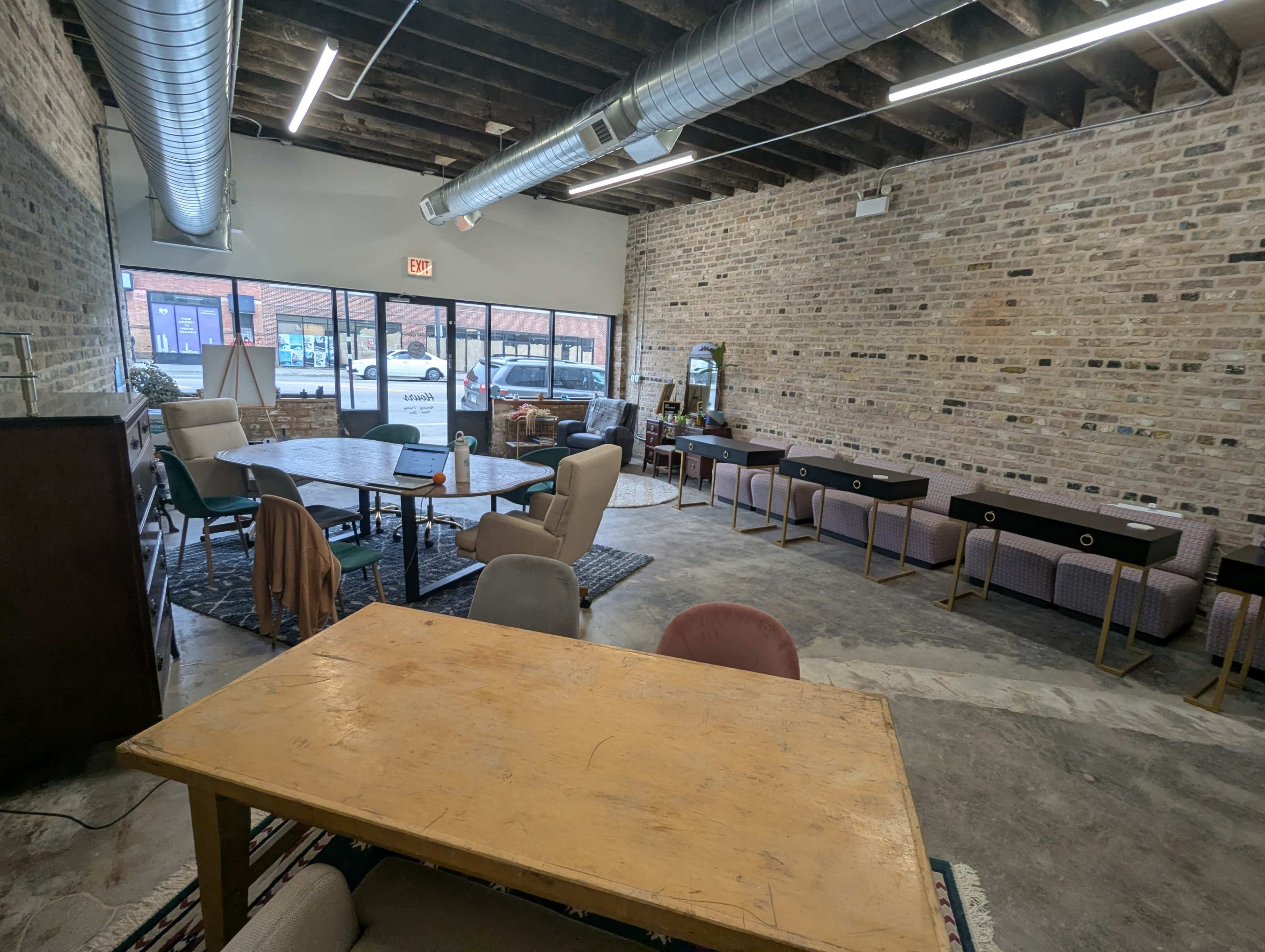 The image shows a spacious, rustic interior with exposed brick walls, a mix of tables and chairs, and large windows allowing natural light to enter.