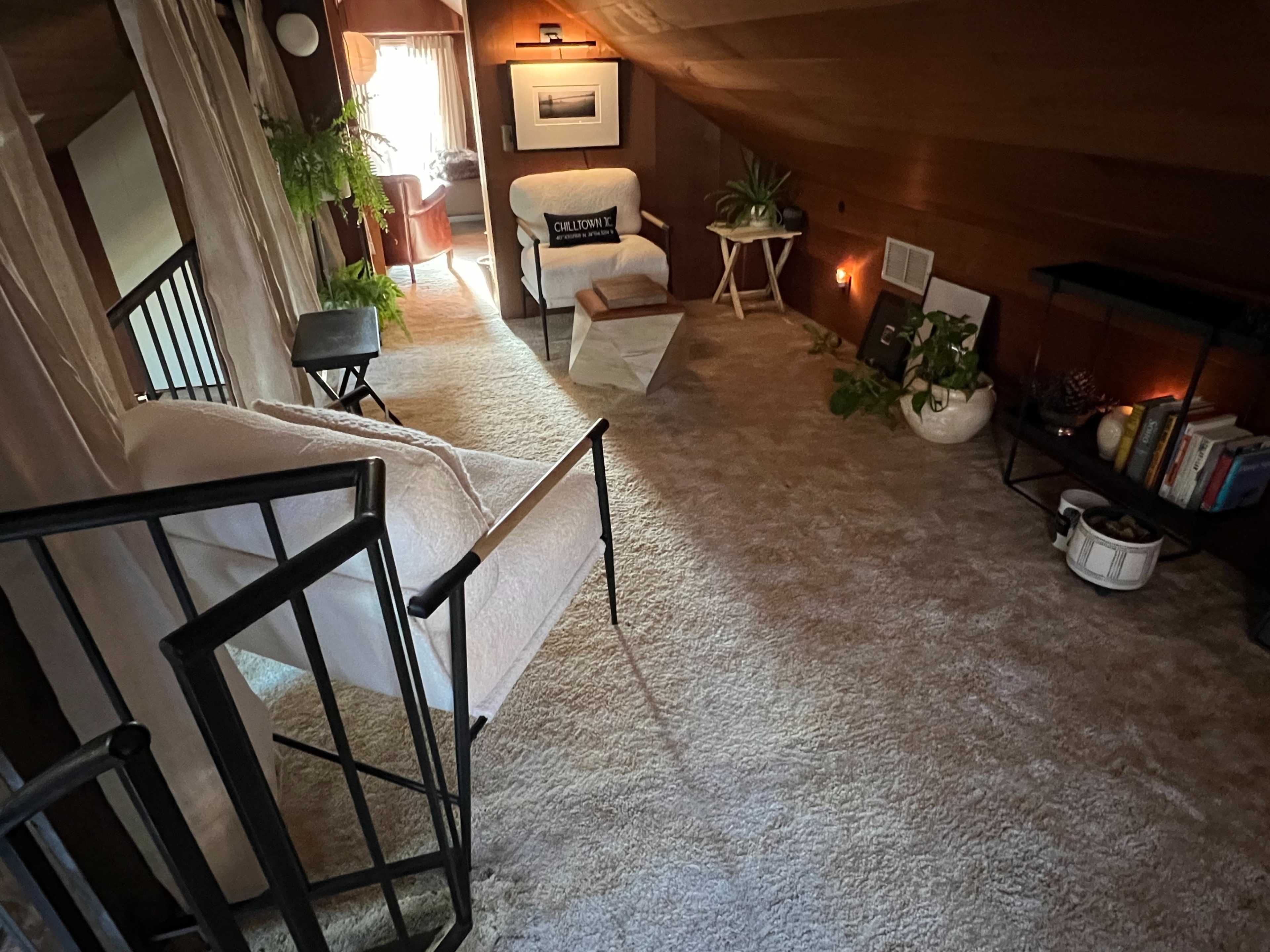 The image shows a cozy, well-lit loft space with a seating area, plants, and decorative elements against a wooden wall.