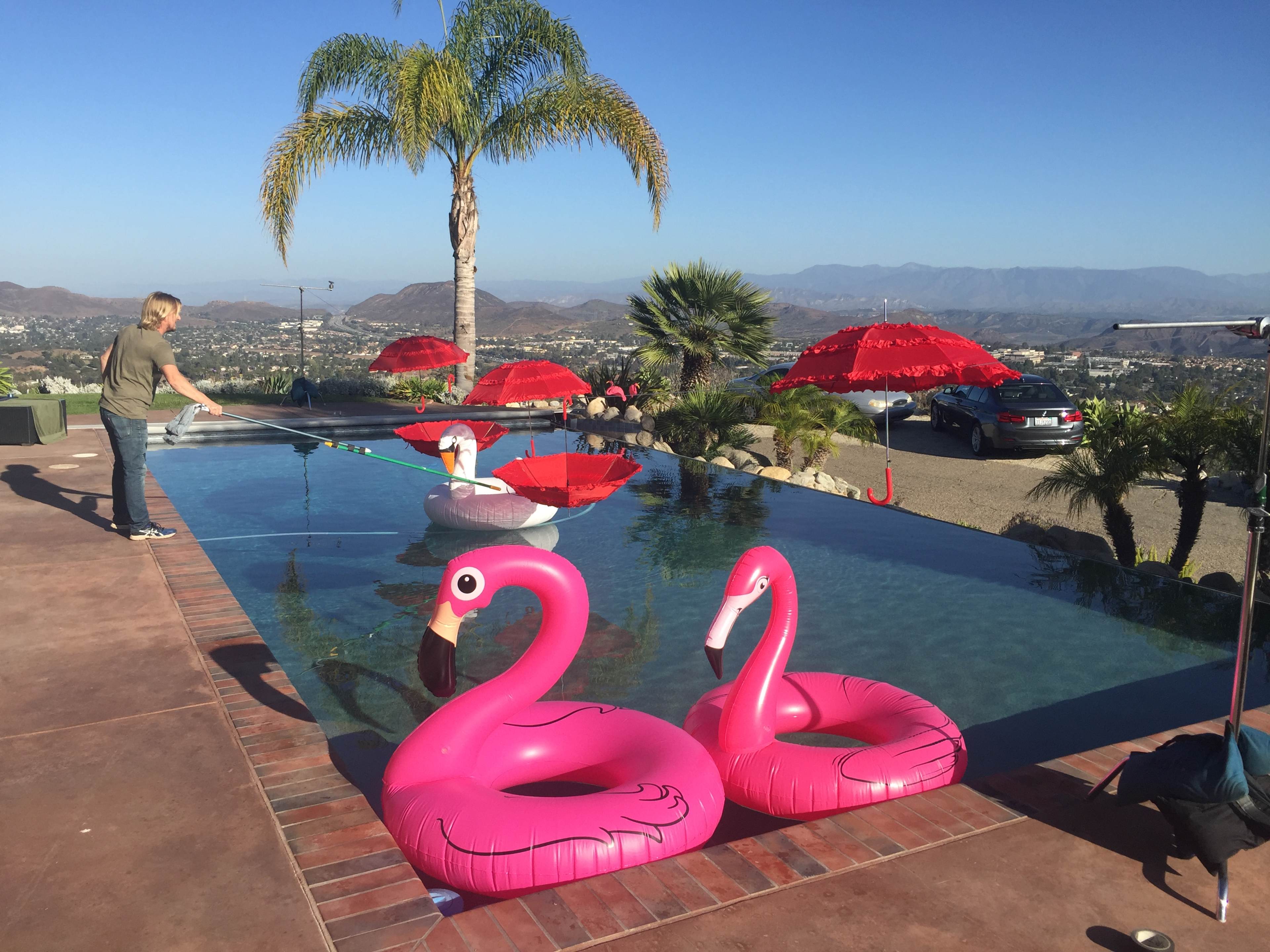 A person stands by a pool with pink flamingo floaties and red umbrellas, overlooking a mountainous landscape.