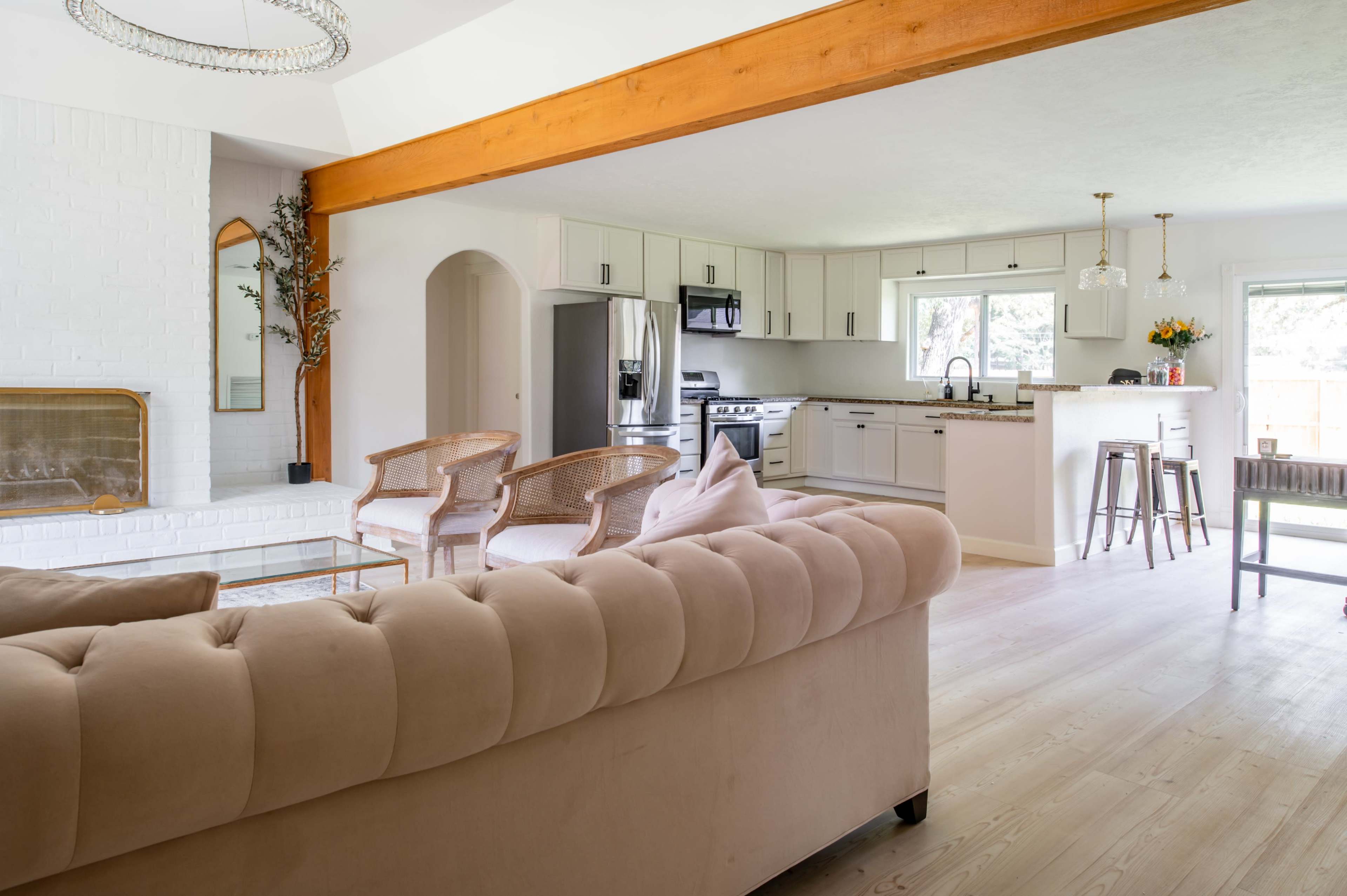A modern living space with a light-colored tufted sofa, a wood beam overhead, and an open kitchen in the background featuring stainless steel appliances and a bar area.