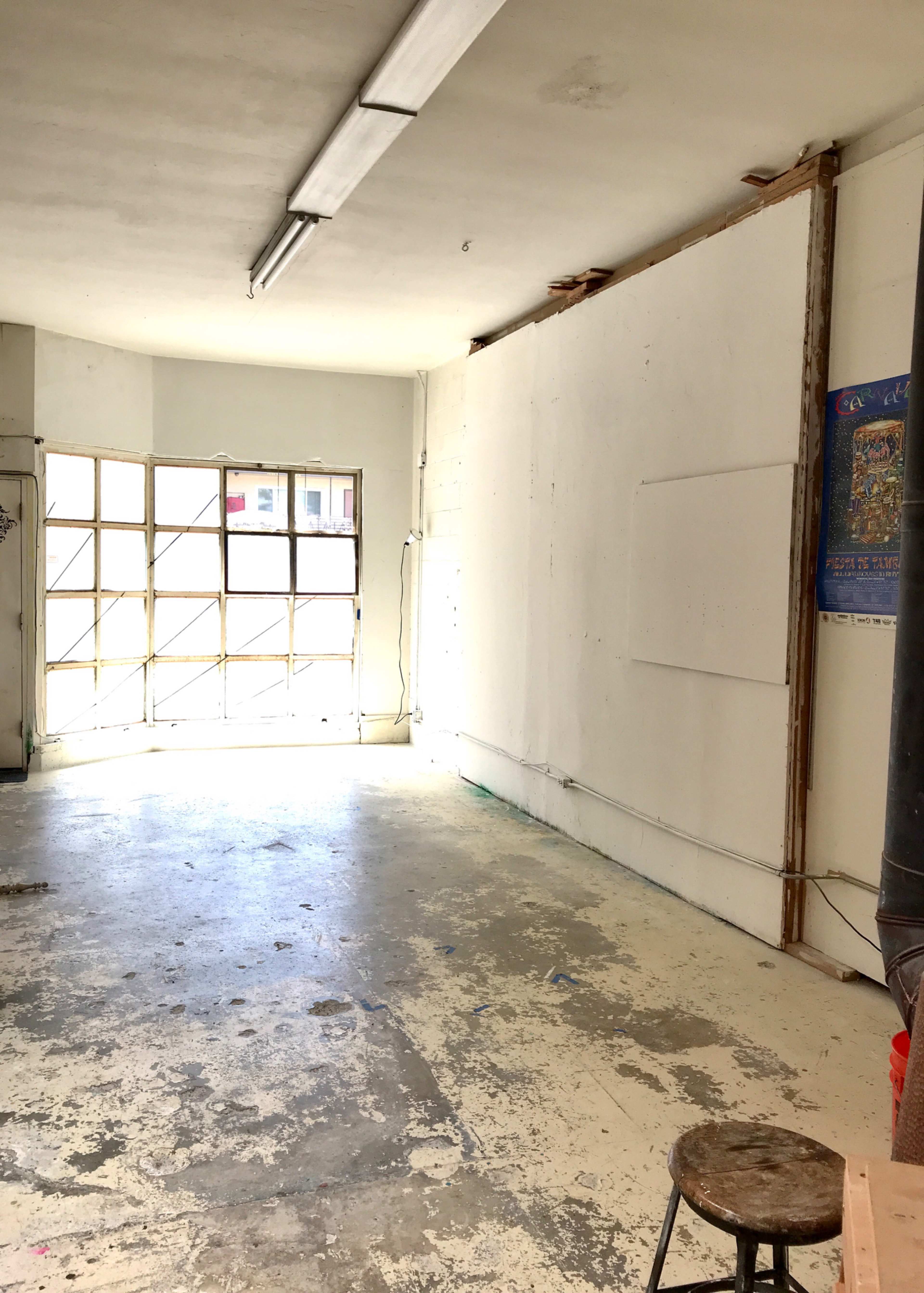 The image shows an empty room with a concrete floor, a large window letting in light, and bare white walls.