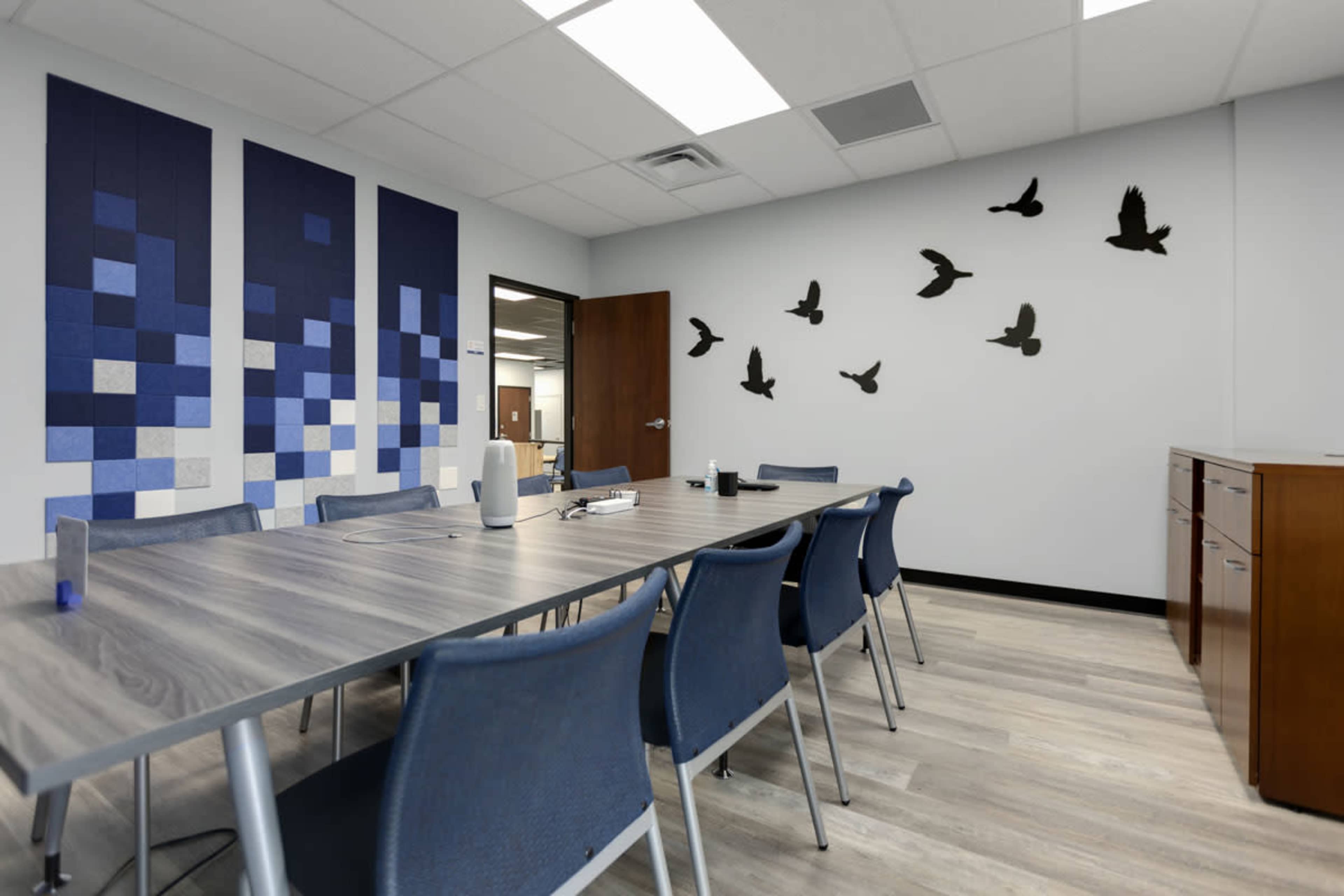 A conference room features a long table surrounded by chairs, with blue and gray wall art and silhouettes of flying birds.