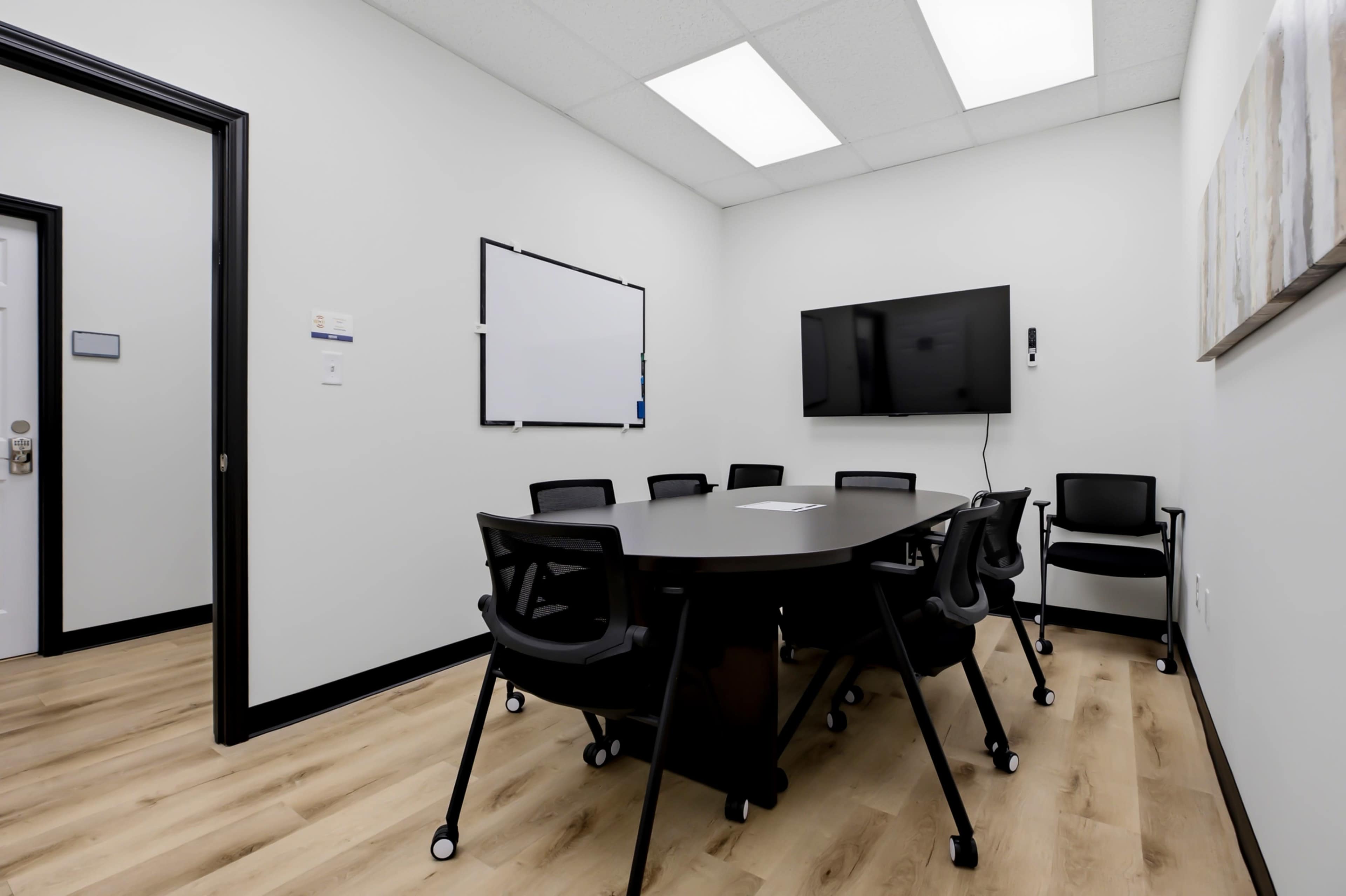 The image shows a conference room equipped with a large oval table, eight black chairs, a wall-mounted television, and a whiteboard.