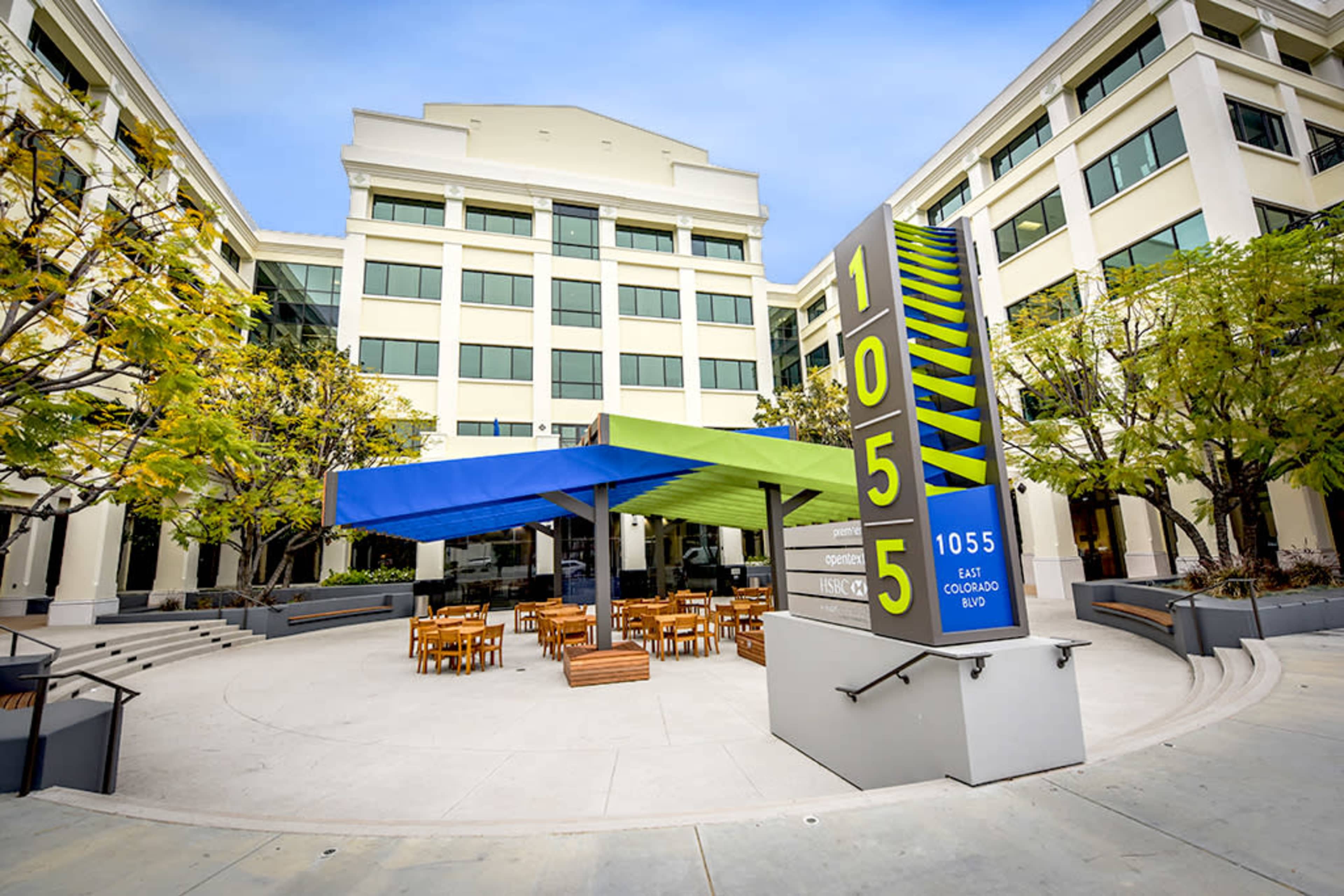 A modern building with a colorful outdoor seating area, featuring a large sign that reads "1055" and a shaded structure over wooden tables and chairs.