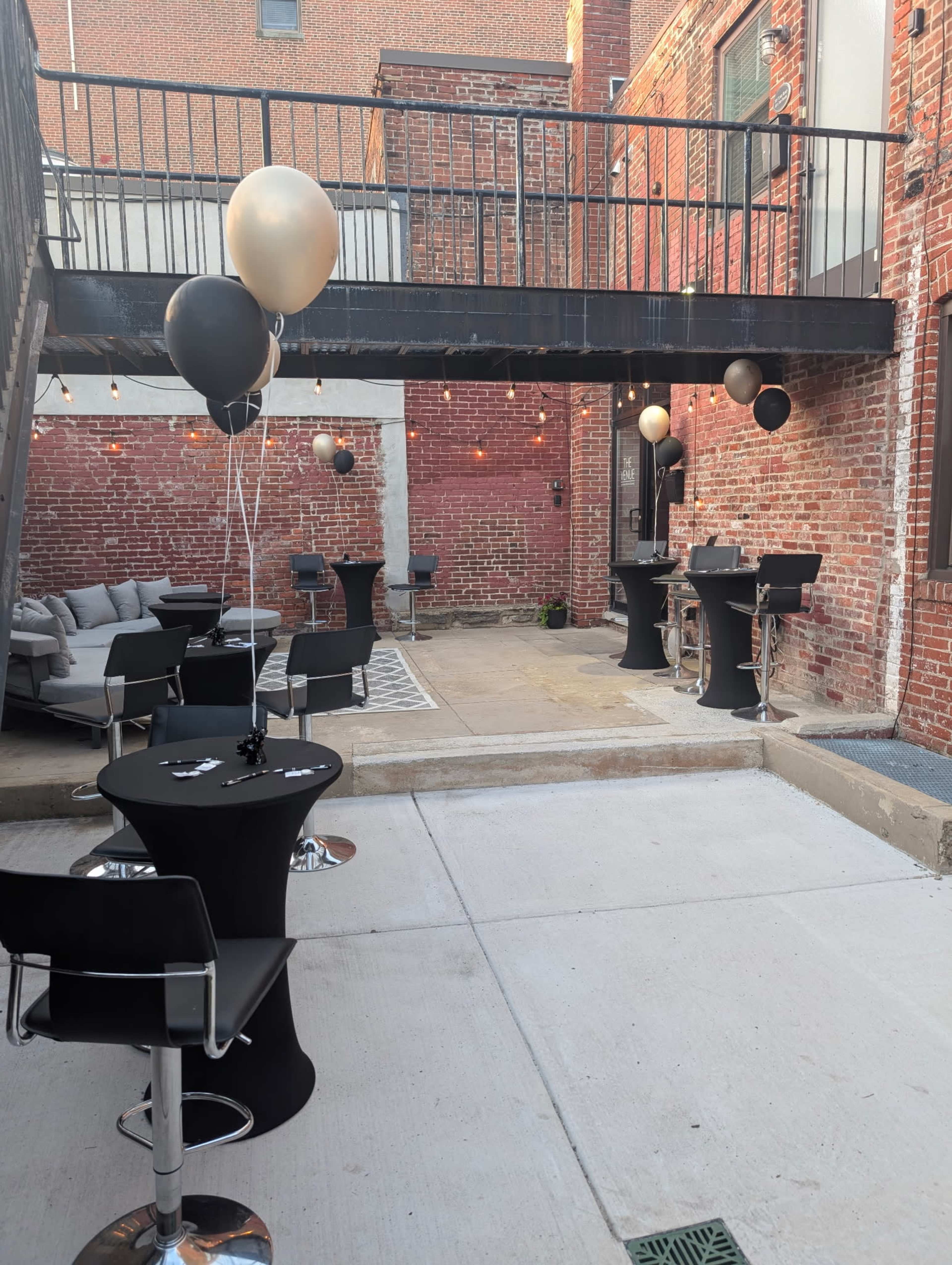 The image shows an outdoor event space with black and gold balloons, several tables, and seating arranged around a brick wall backdrop.