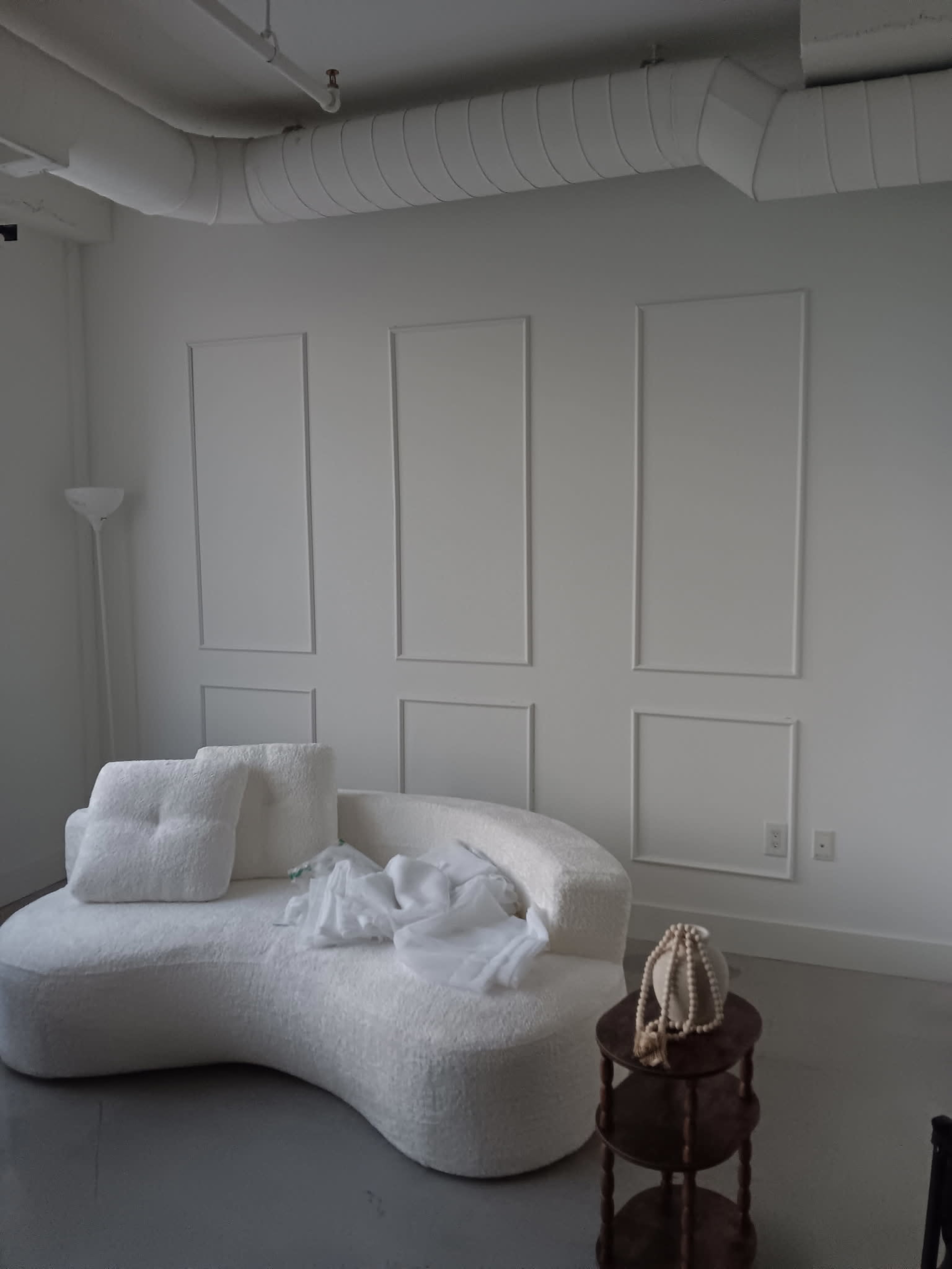The image shows a modern, curved white sofa placed against a wall with decorative paneling, alongside a small side table and a lamp.