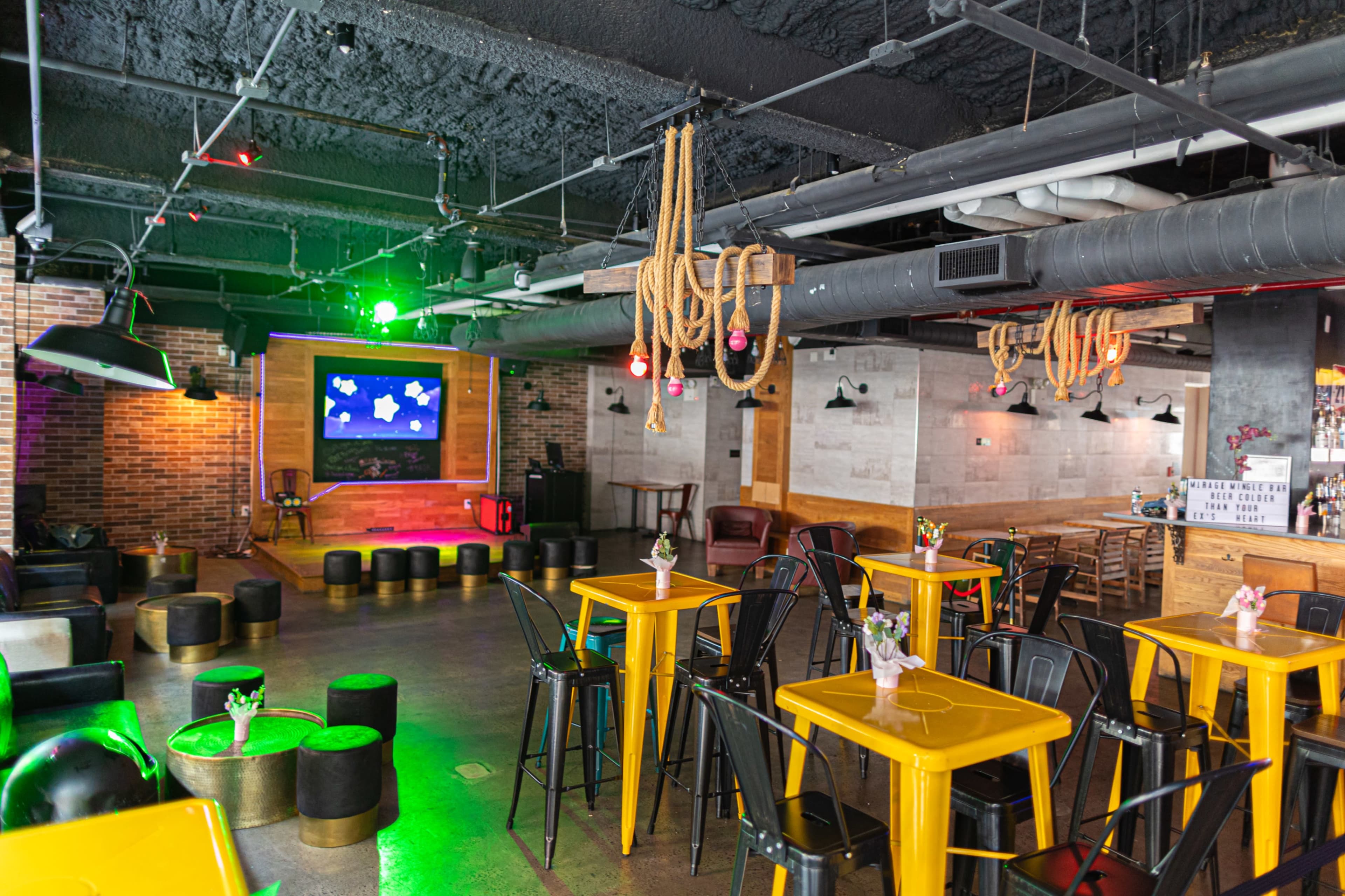 A spacious lounge area features yellow tables, black seating, and a stage with colorful lighting.