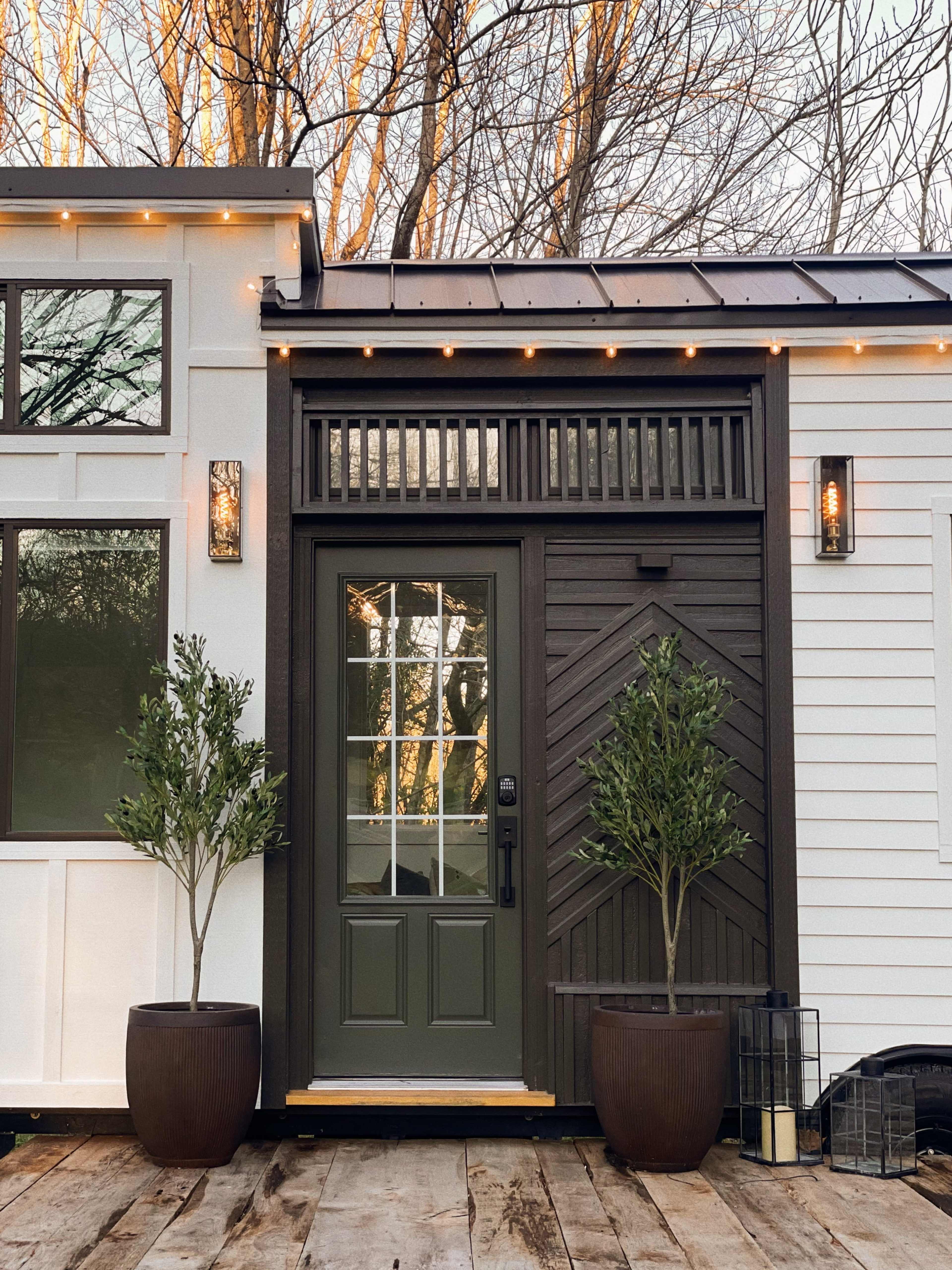 A green front door is flanked by two potted plants on a wooden deck, with string lights above and trees in the background.