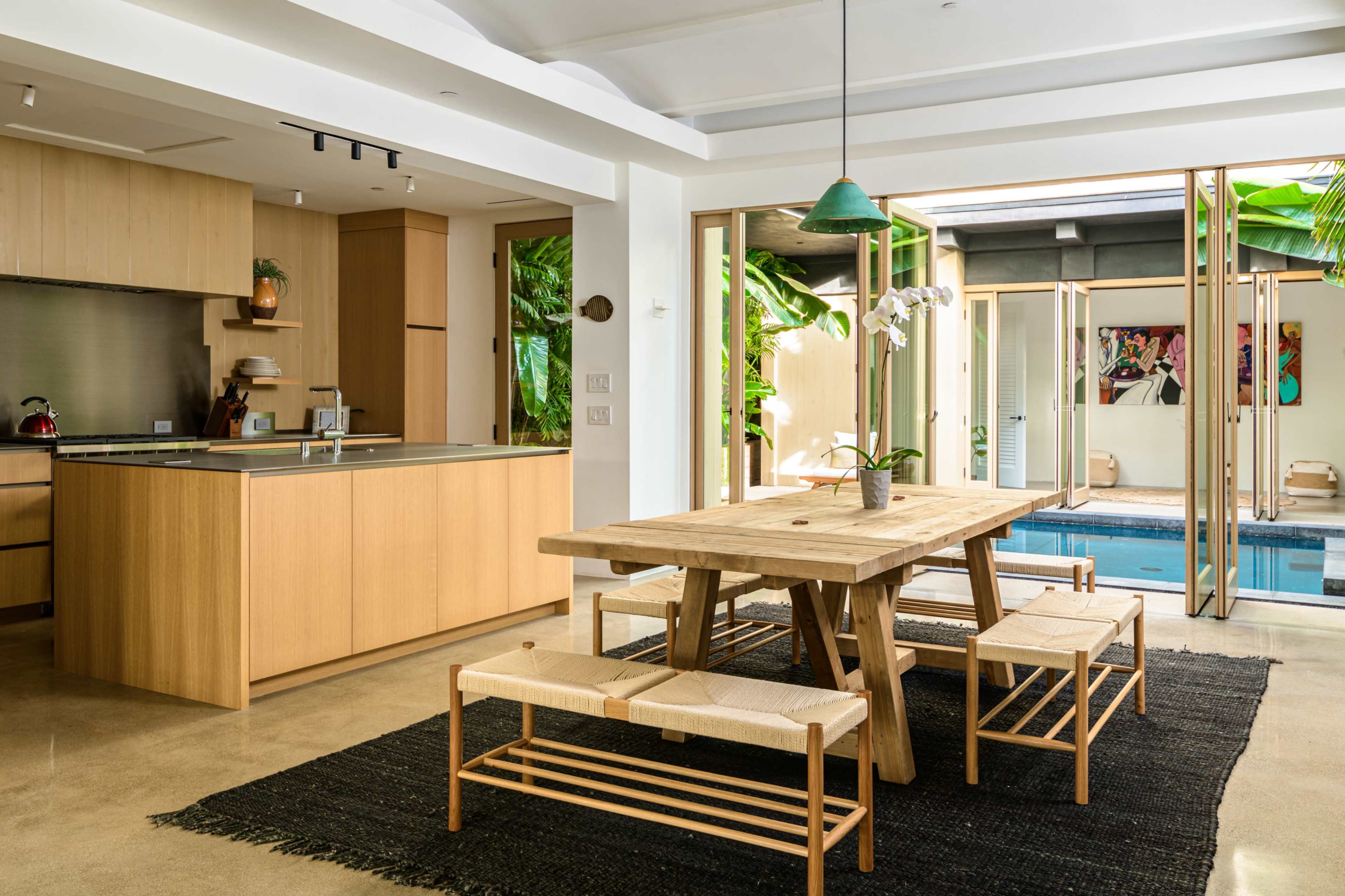 A modern dining area featuring a wooden table and benches, with a kitchen in the background and large sliding doors opening to a pool surrounded by greenery.