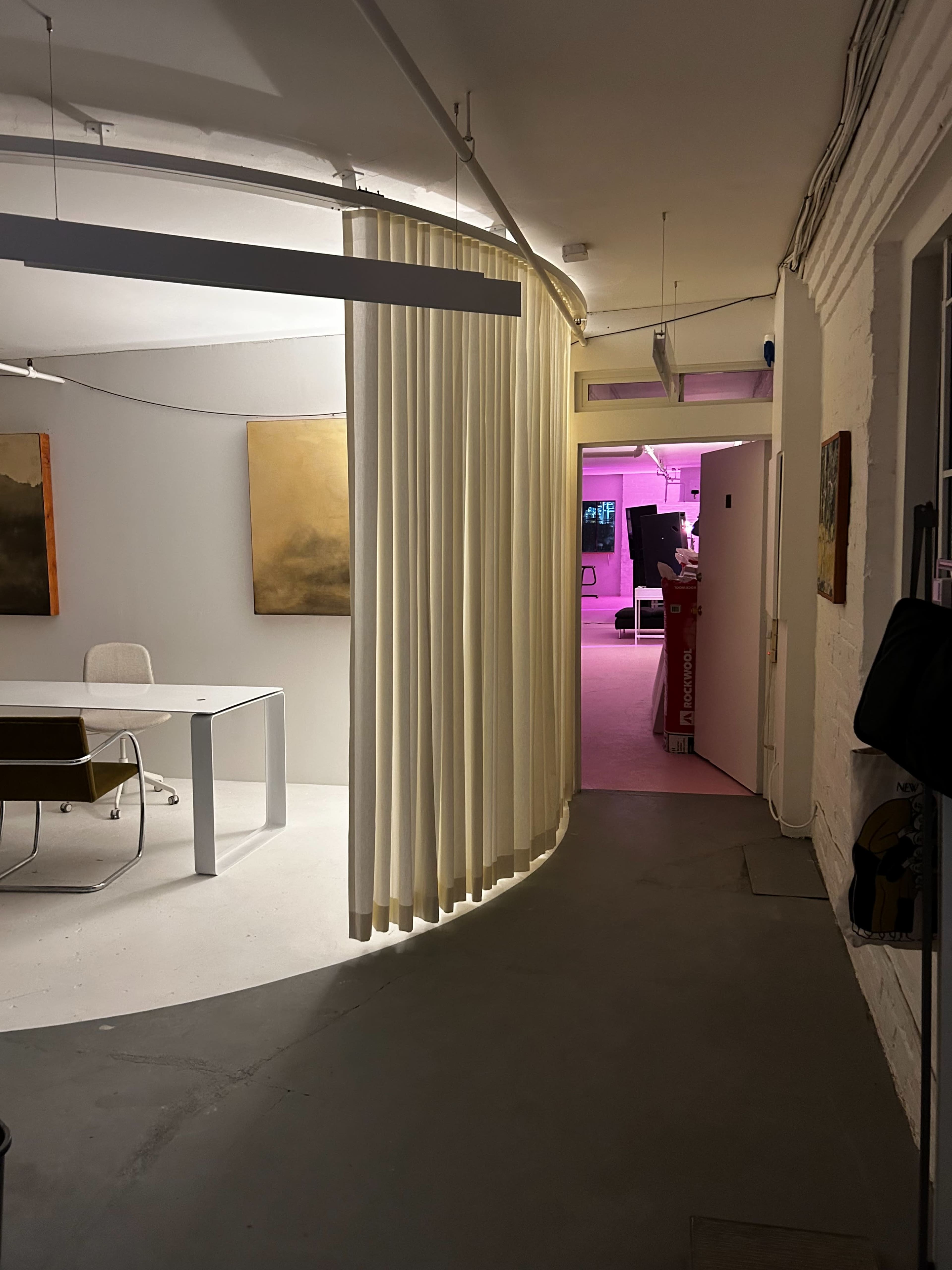 The image shows a modern office space with a curved wall featuring draped curtains, leading to a brightly lit room in pink hues at the far end.