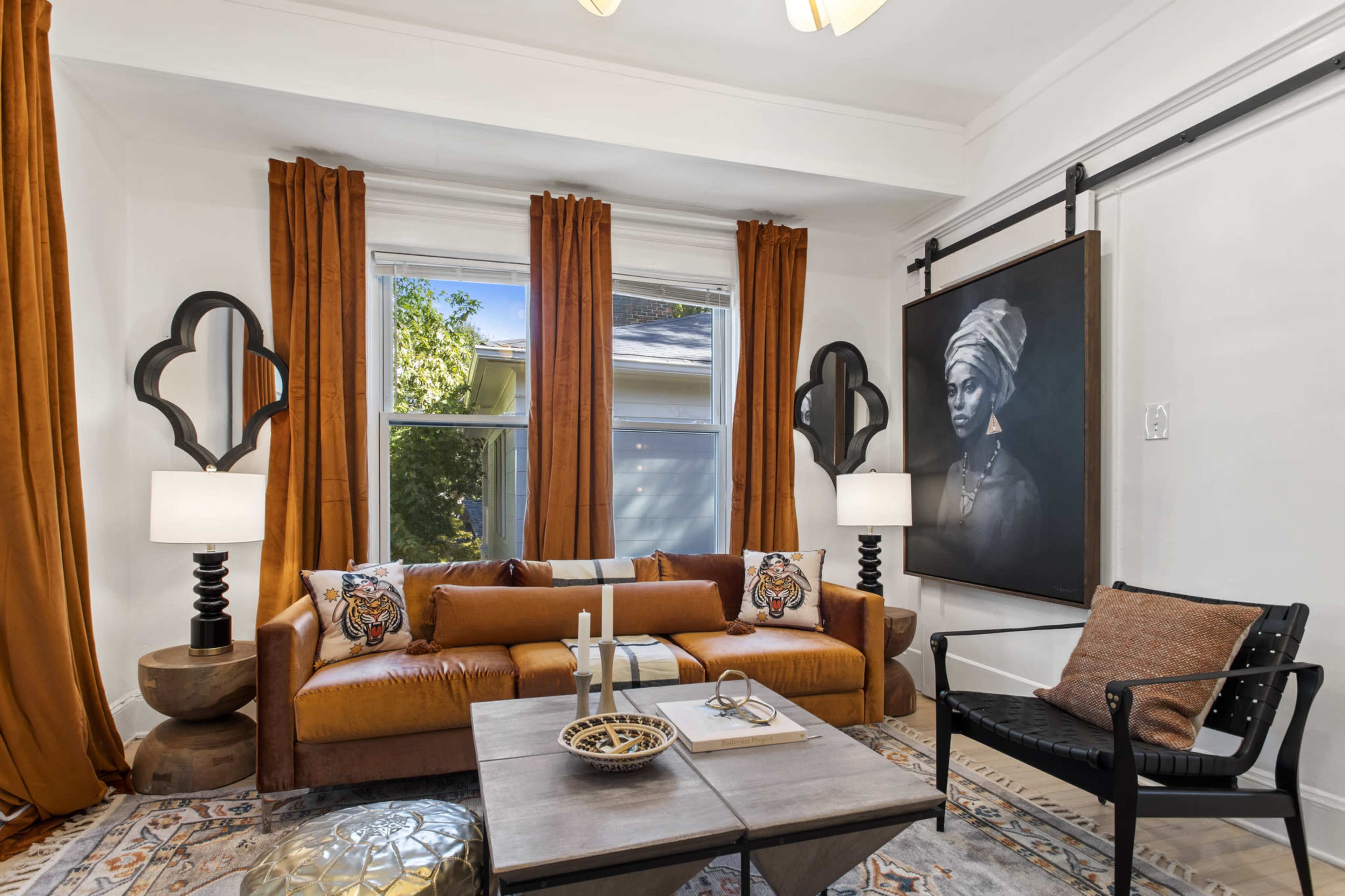 A living room features a brown sofa with decorative pillows, two lamps on side tables, a large portrait on the wall, and warm orange curtains.
