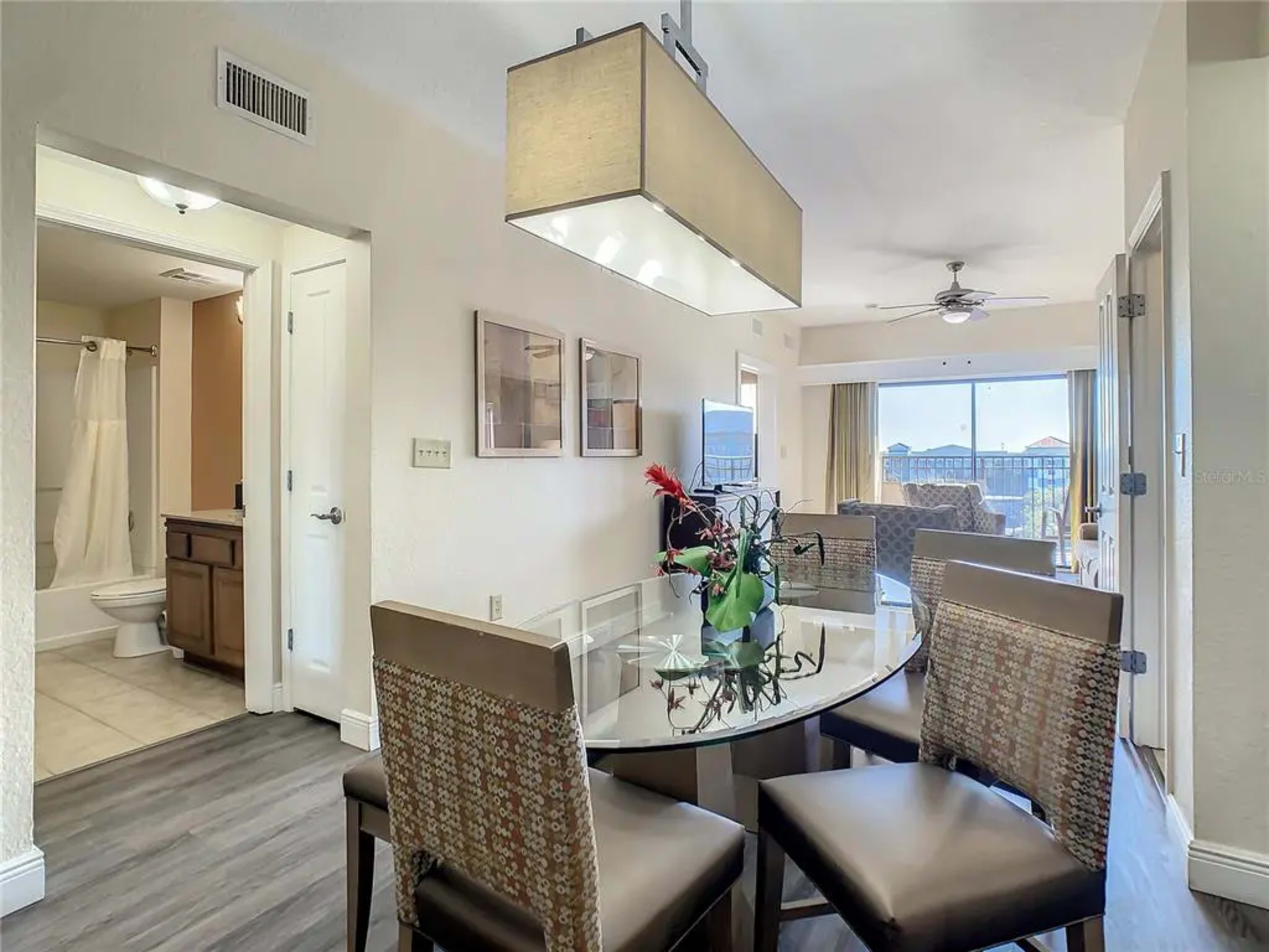 A dining area with a circular glass table and four chairs is situated near an open entryway leading to a bathroom and a living room with large windows.
