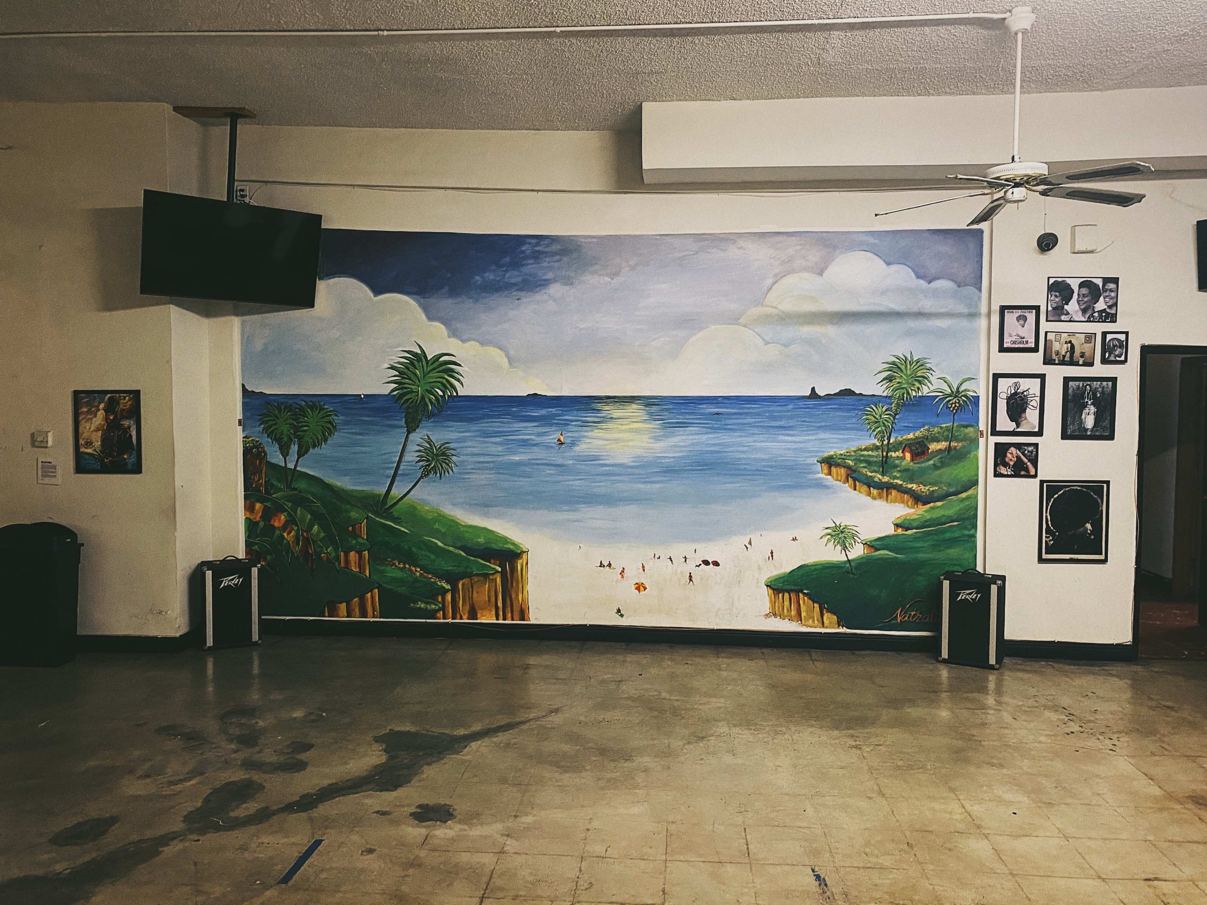 An interior space featuring a large mural of a tropical beach scene on one wall, with palm trees and distant islands, and framed photographs displayed alongside it.