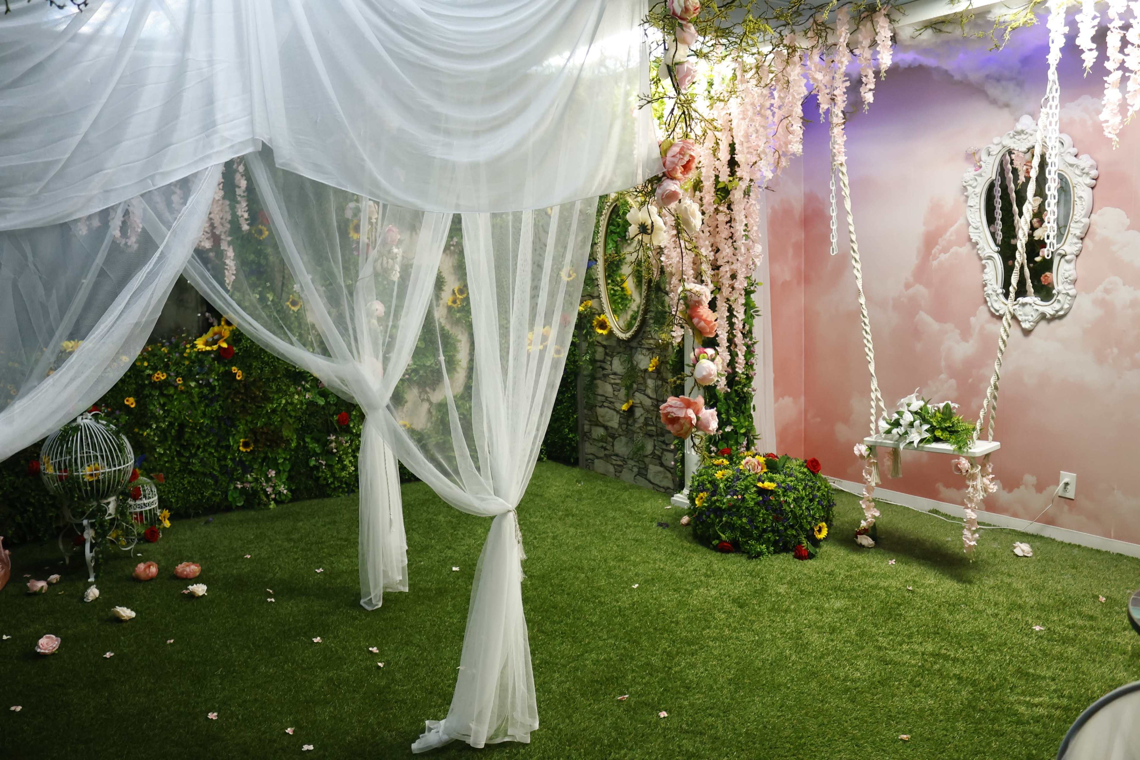 A decorative indoor space features sheer white drapery, artificial flowers, and a grassy floor.