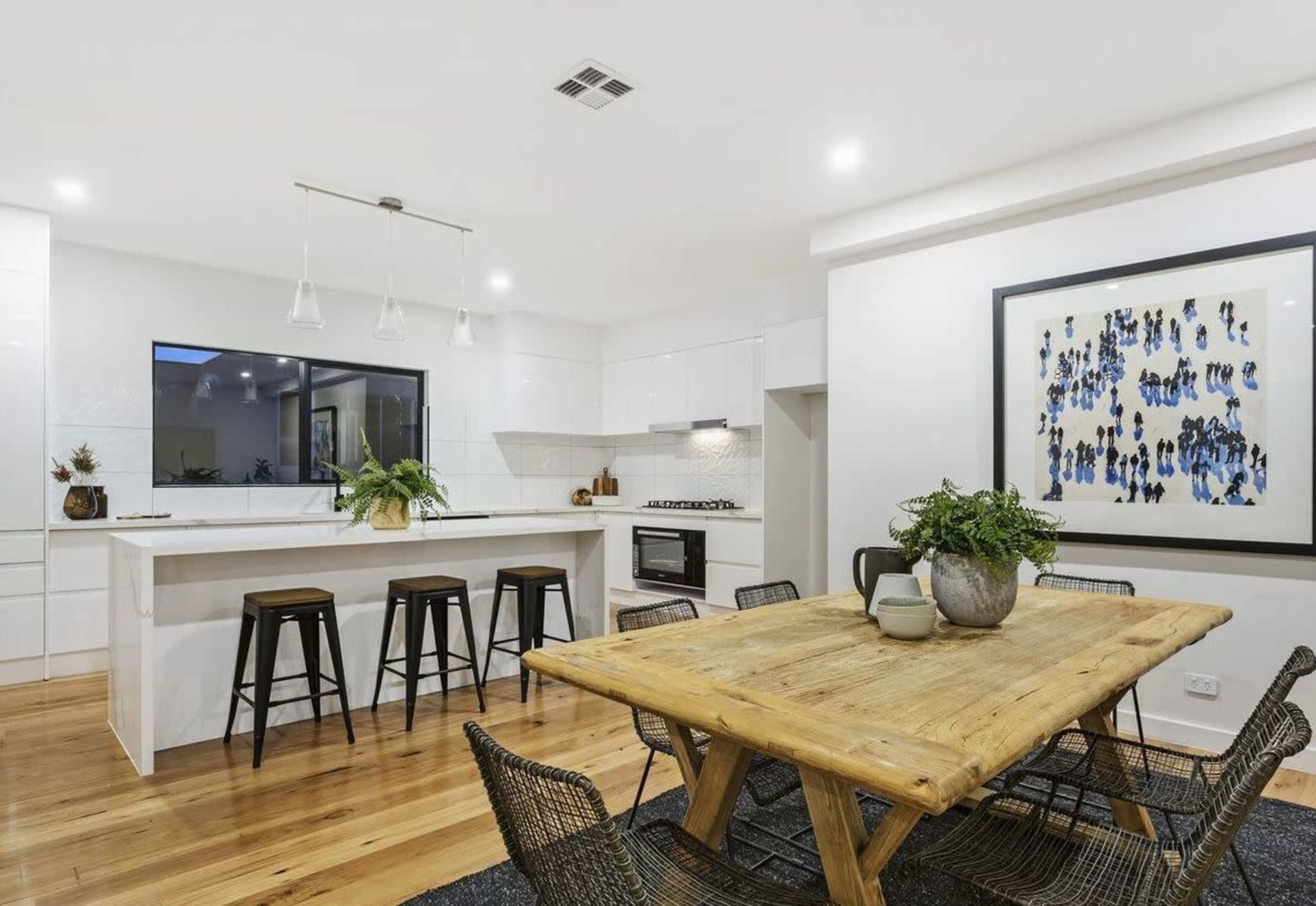 A modern kitchen features a wooden dining table surrounded by wire chairs, with a kitchen island and sleek cabinetry in the background.