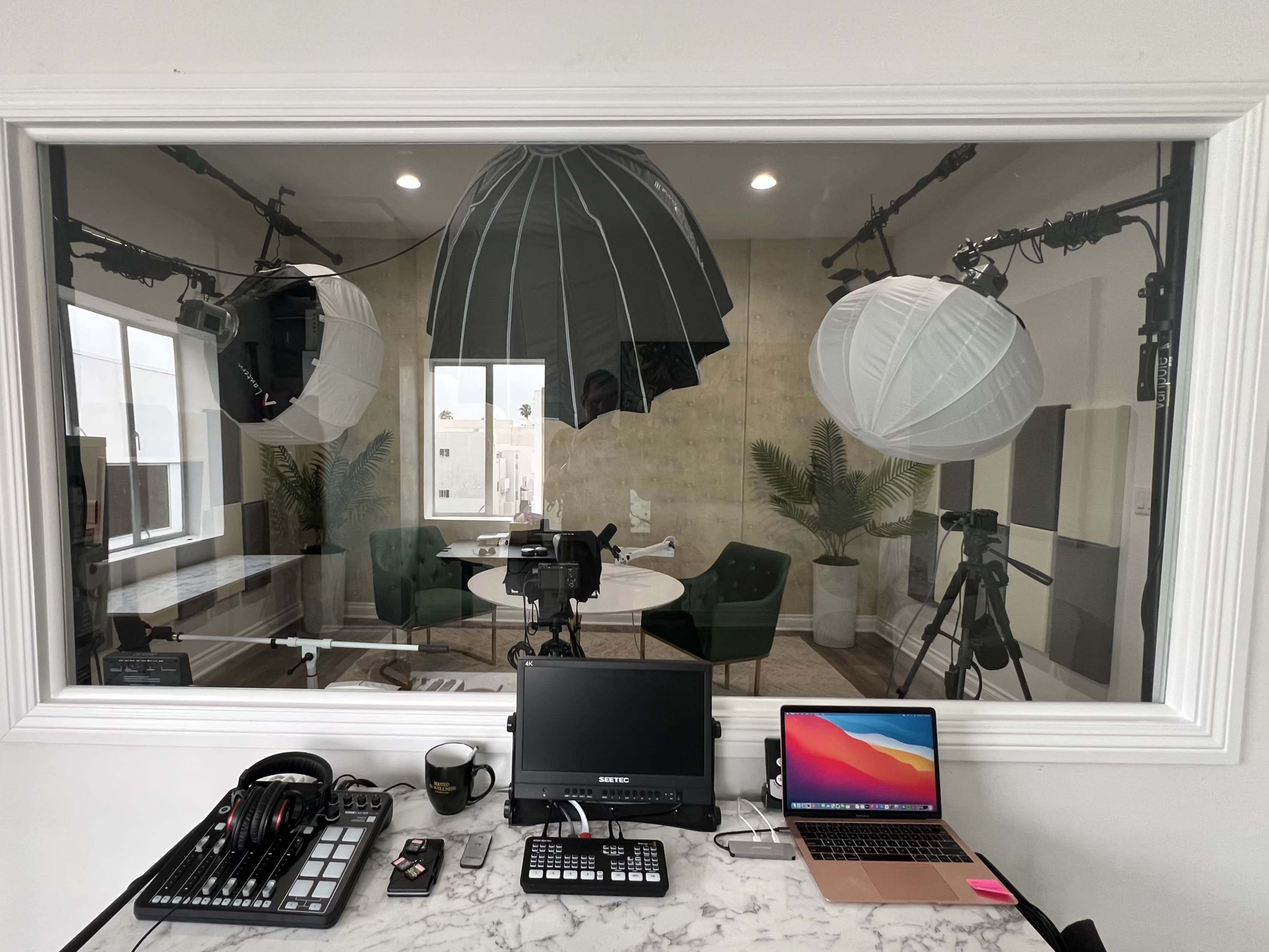 A well-lit studio setup is visible through a large glass window, featuring cameras, lighting equipment, and a workspace with a laptop and audio gear.